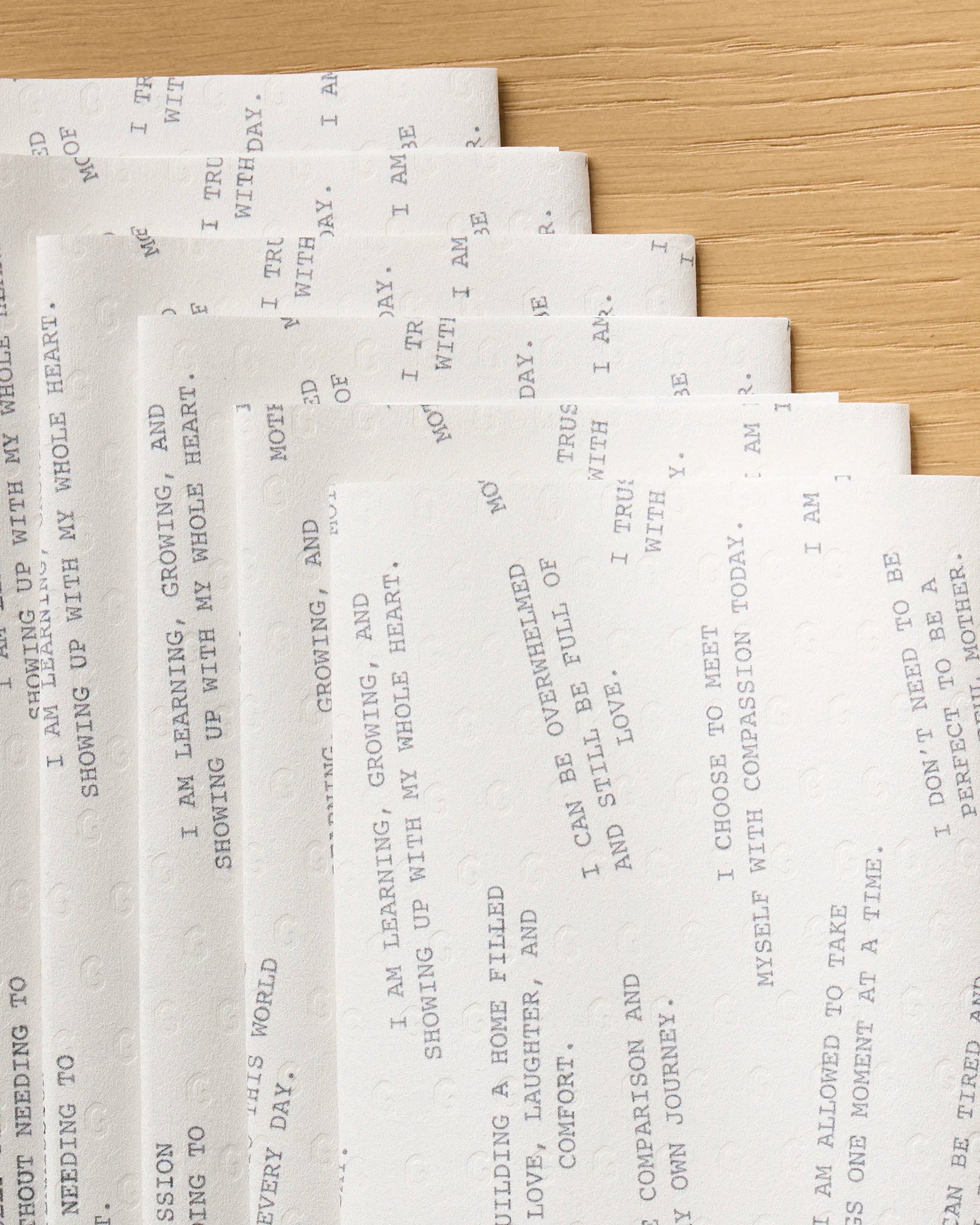 Eight sheets of Geometry’s “Reminders for Mom” eco-friendly white paper towels with faint printed text are fanned out on a wooden surface, their corners neatly aligned. The light print makes the reminders difficult to read.