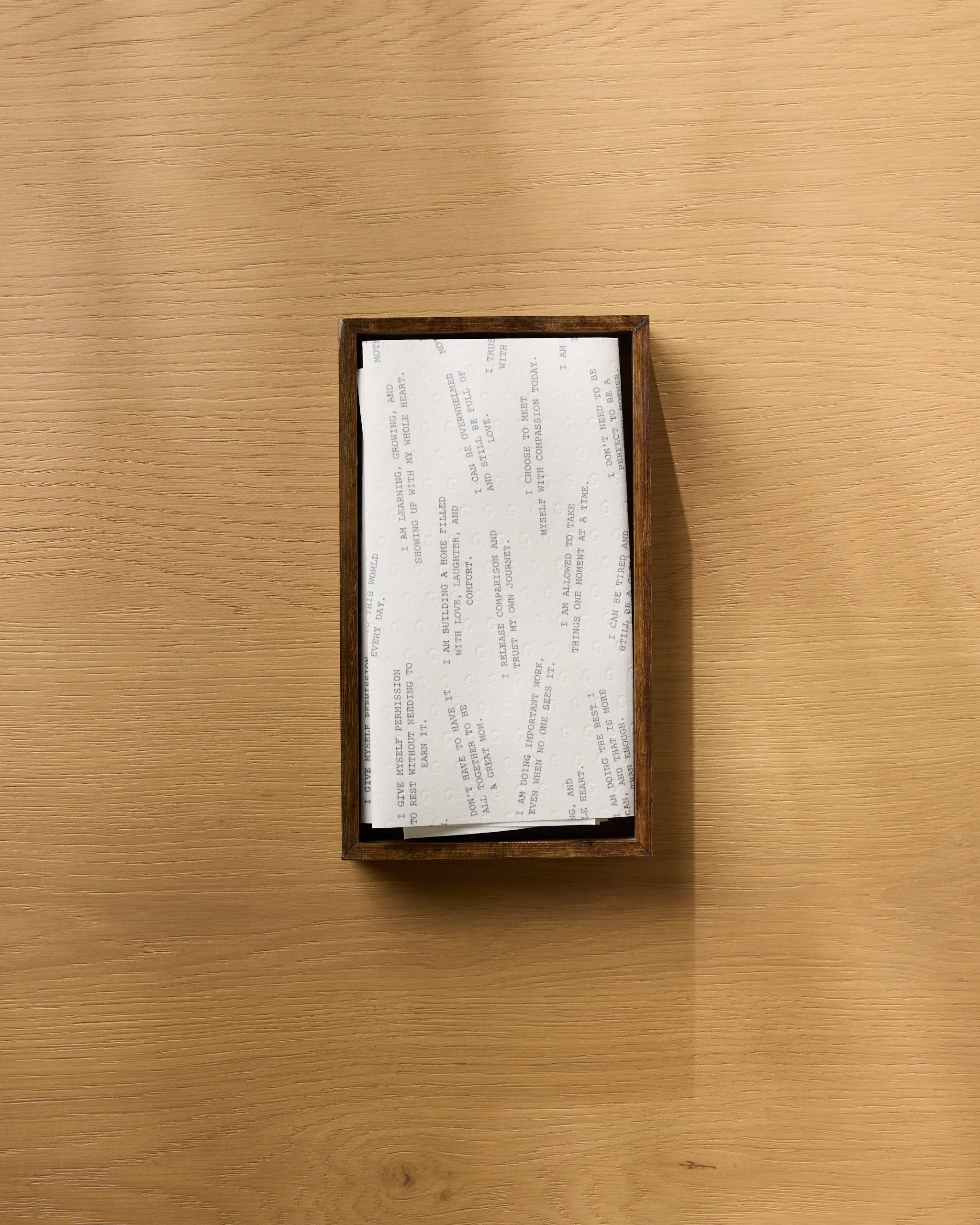 The Reminders for Mom box by Geometry sits on a light wood surface, filled with recycled paper towels and randomly arranged white note slips featuring text, as seen from above.