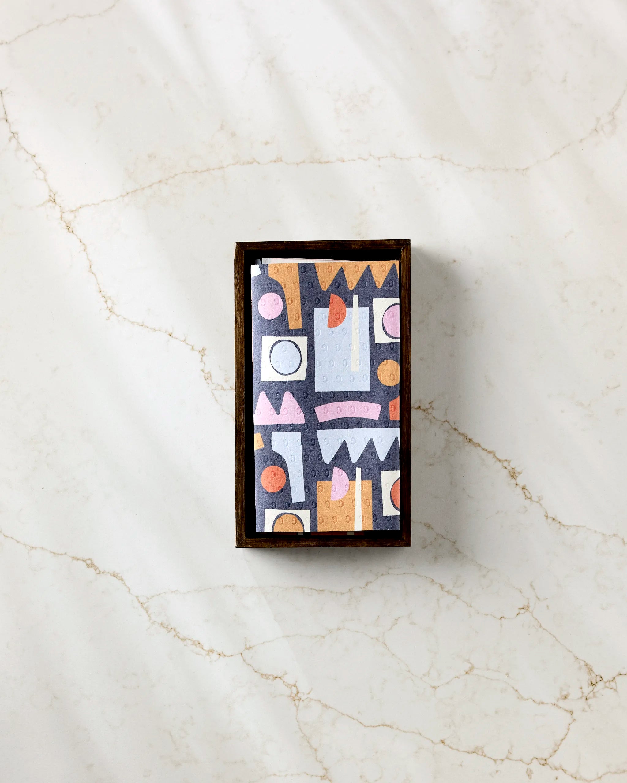 A rectangular box with a colorful geometric pattern sits on marble. Inside, Claire Iglesias' Retro Shapes Fiesta offers eco-friendly kitchen towels—reusable paper towel alternatives in pastel pink, orange, gray, and black.