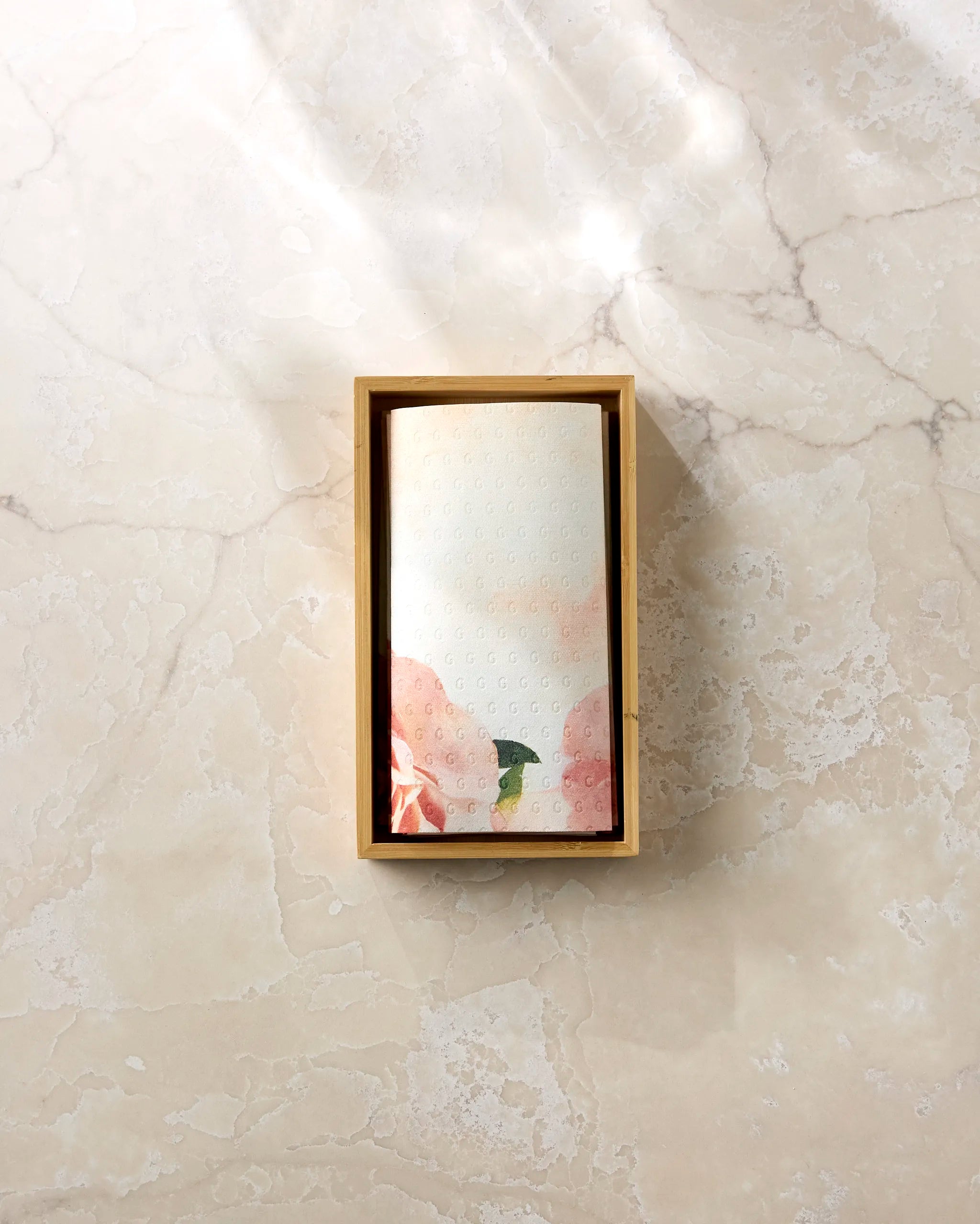 A single roll of Stefanie Bales’ Rhapsody in Bloom decorative recycled paper towels, featuring floral and landscape designs, rests neatly in a wooden box on a light marble surface.