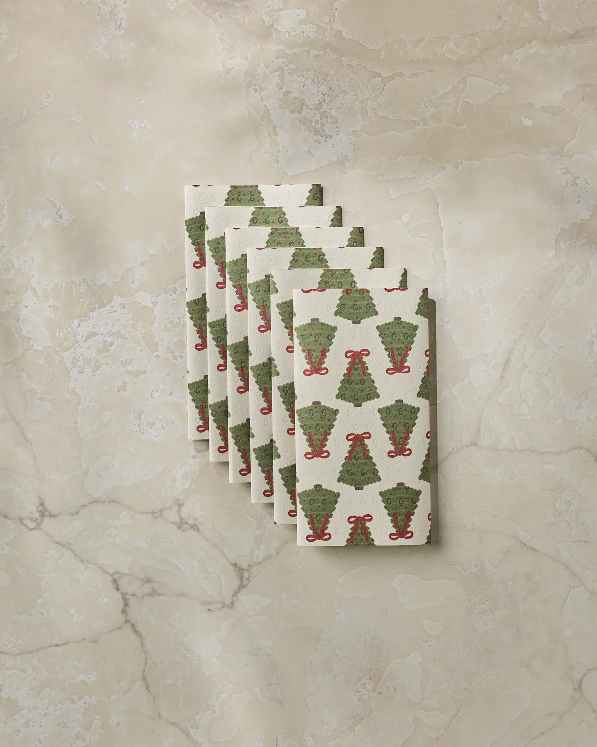 Six “Ribbon Pines” holiday-themed napkins from Geometry, featuring green Christmas trees and red bows, are fanned out on a beige marble surface—offering an eco-friendly alternative to disposable paper towels for your festive gatherings.