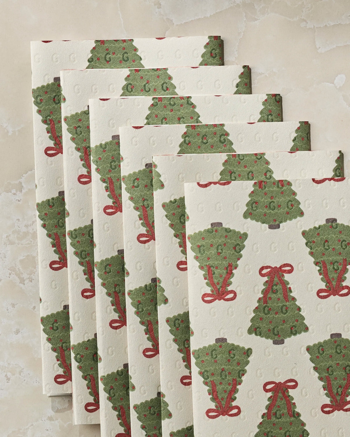 Five “Ribbon Pines” holiday-themed napkins from Geometry, featuring green Christmas trees adorned with red bows and ornaments, are arranged in a fanned-out pattern on a marble surface—offering an eco-friendly alternative to disposable paper towels for your festive gatherings.