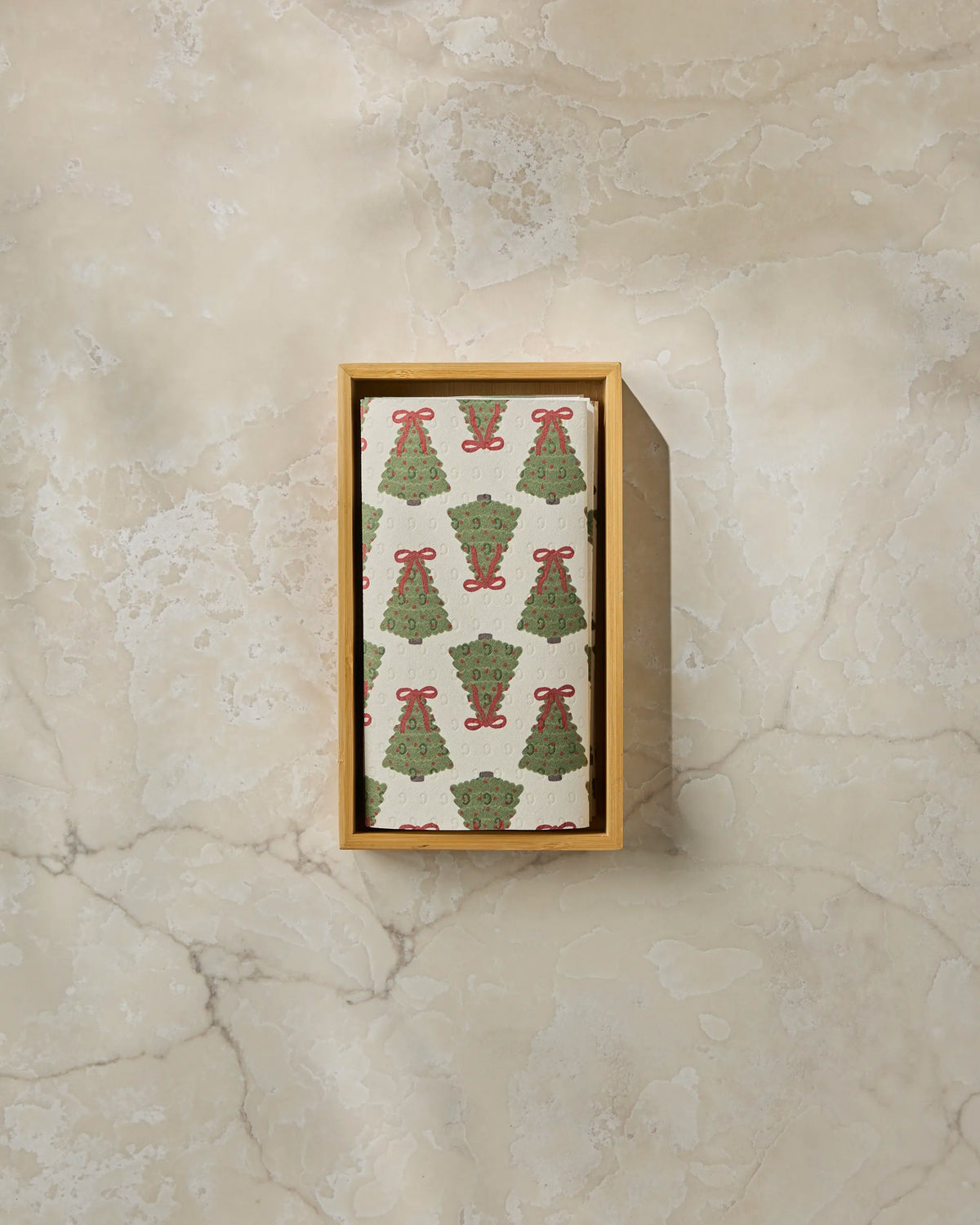 The “Ribbon Pines” cloth napkin by Geometry, featuring a pattern of green Christmas trees and red bows, is elegantly arranged in a rectangular wooden box on a beige marble surface, presenting a festive and eco-friendly alternative to disposable paper towels.