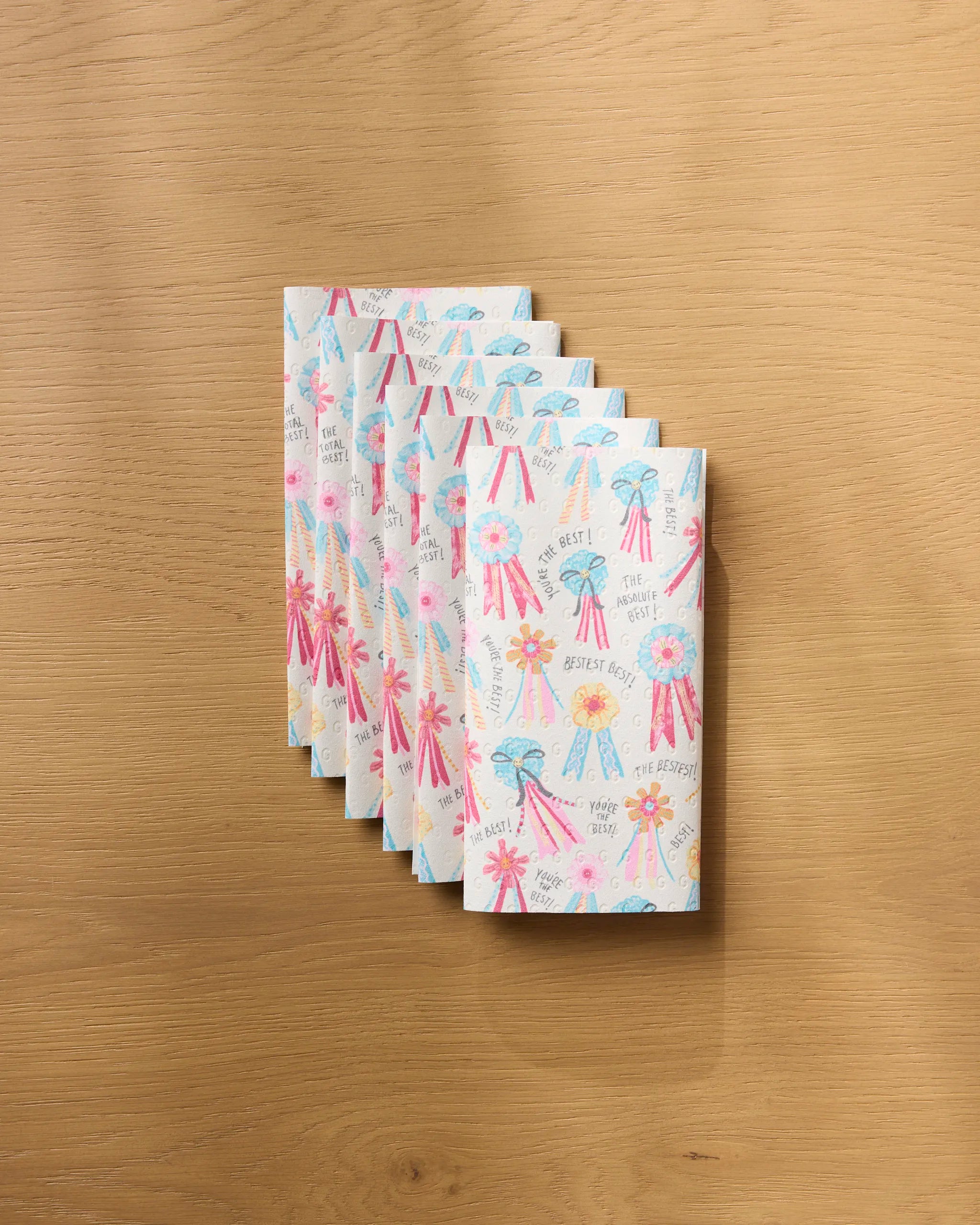 Seven “Rosettes for You” greeting cards by Geometry, featuring colorful ribbon designs and messages like Best Boy, The Best, and Very Sweet, are neatly fanned out on an eco-friendly wooden surface.