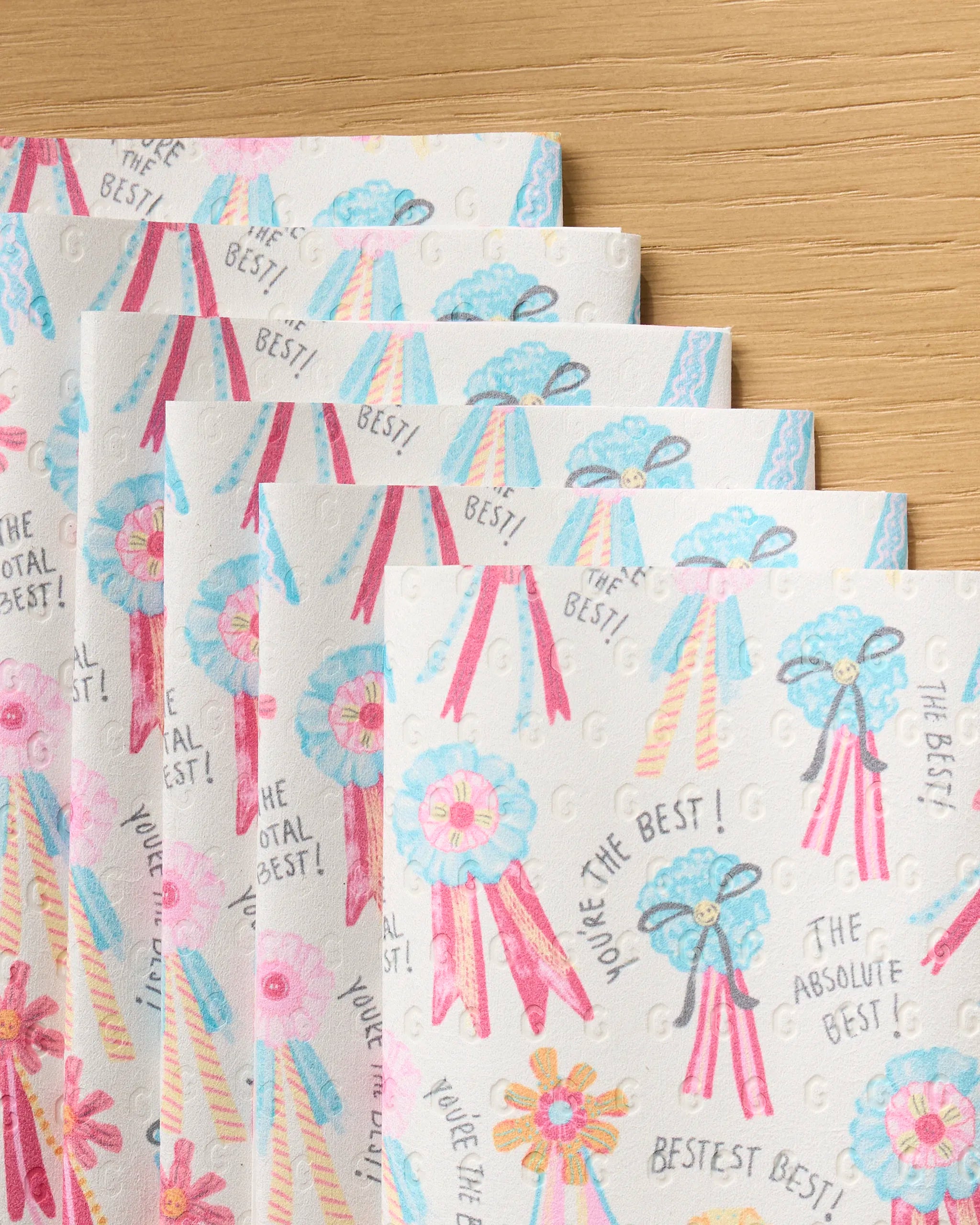 Several “Rosettes for You” sheets by Geometry, featuring colorful award ribbon designs and phrases like “You’re the Best!”, are fanned out on a wooden surface—try their reusable paper towels for an eco-friendly craft option.