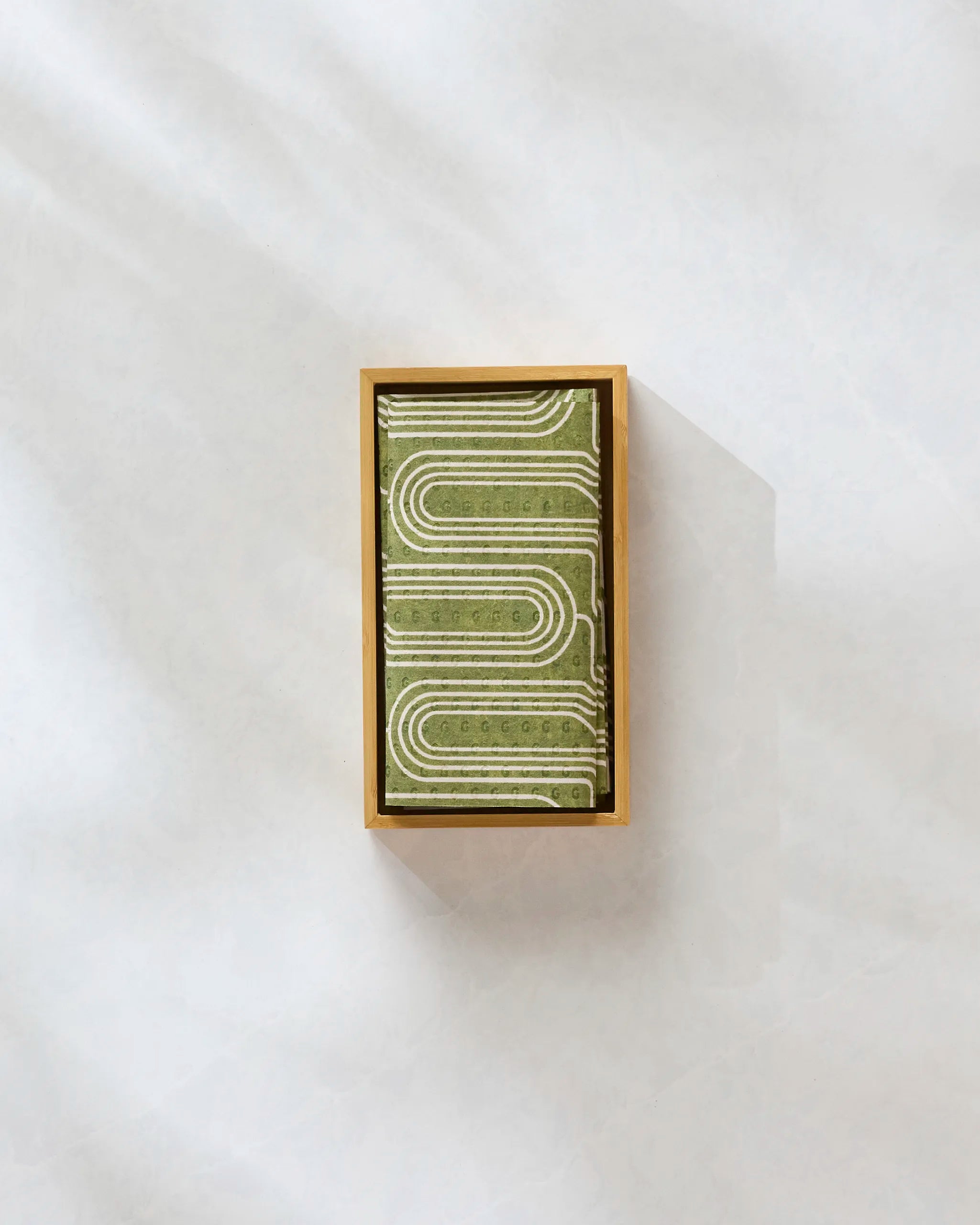 The Roslyn bar by Amy MacCready is a rectangular matcha green chocolate in a gold box with a geometric pattern of white lines, displayed on a light, textured surface resembling eco-friendly paper towels.