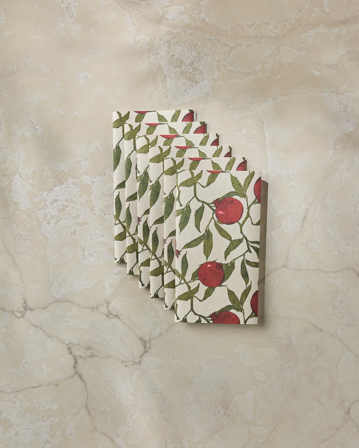 Six folded Ruby Rind napkins by Geometry—featuring a red and green pomegranate and leaf pattern, and crafted from environmentally friendly recycled materials—are arranged in a fan shape on a beige marble surface.