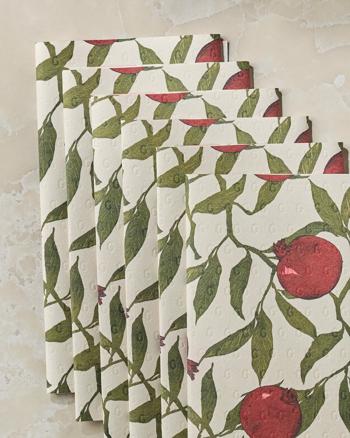 Five Ruby Rind paper napkins from Geometry, made from recycled materials, are fanned out on a light marble surface, each adorned with a pattern of green leaves and red pomegranates.