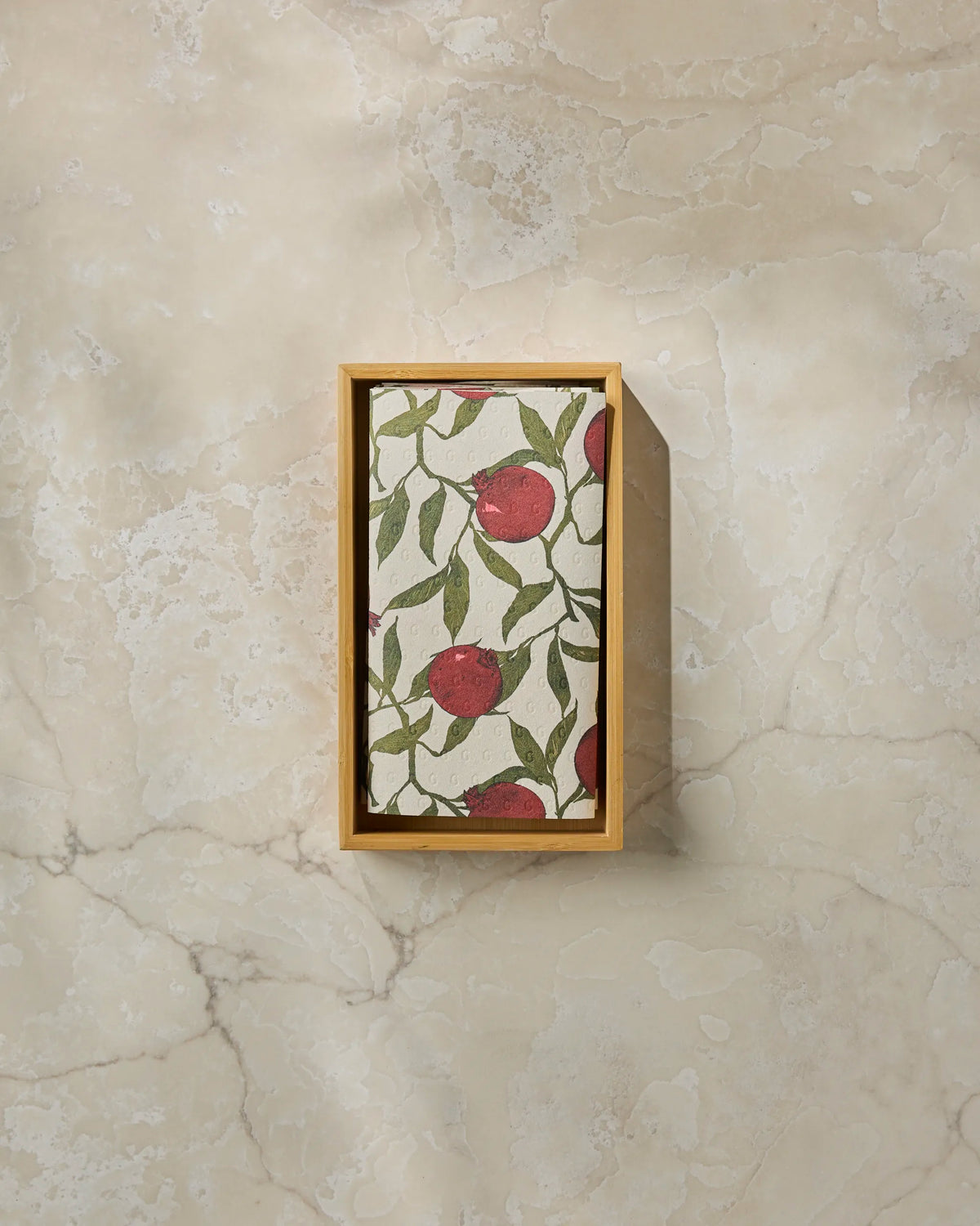 A rectangular wooden tray holds Geometry’s “Ruby Rind” eco-friendly patterned cloth, featuring vibrant red pomegranates and green leaves, displayed on a light marble surface.
