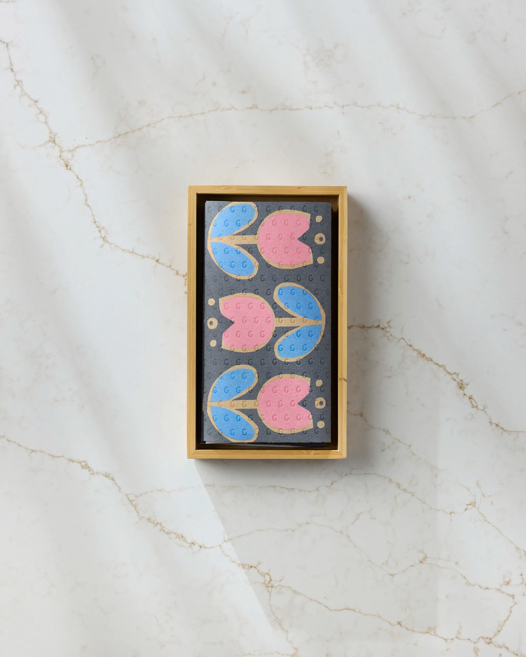 The Scandi Tulips by Geometry is a rectangular wooden box featuring six cookies with pink and blue tulip designs, neatly arranged on black eco-friendly paper towels atop a white marble surface.