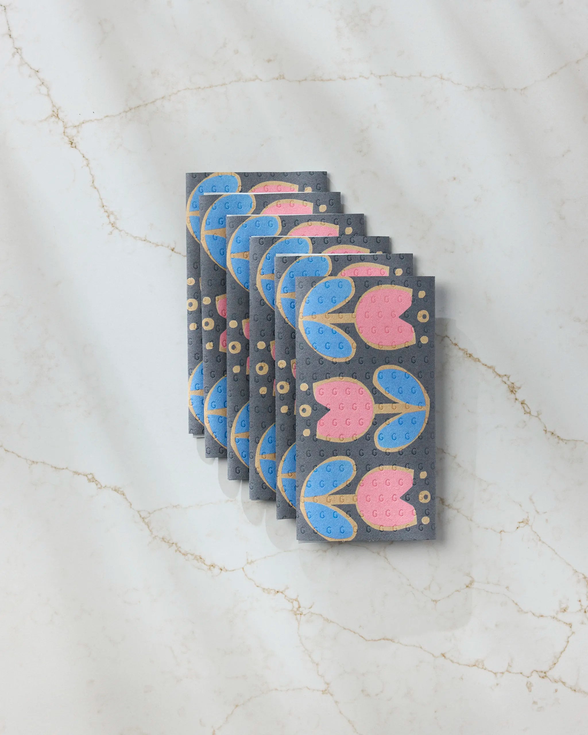 A stack of six Geometry Scandi Tulips reusable paper towels, featuring a blue and pink tulip floral pattern on gray, arranged neatly on a white marble surface with gold veining.