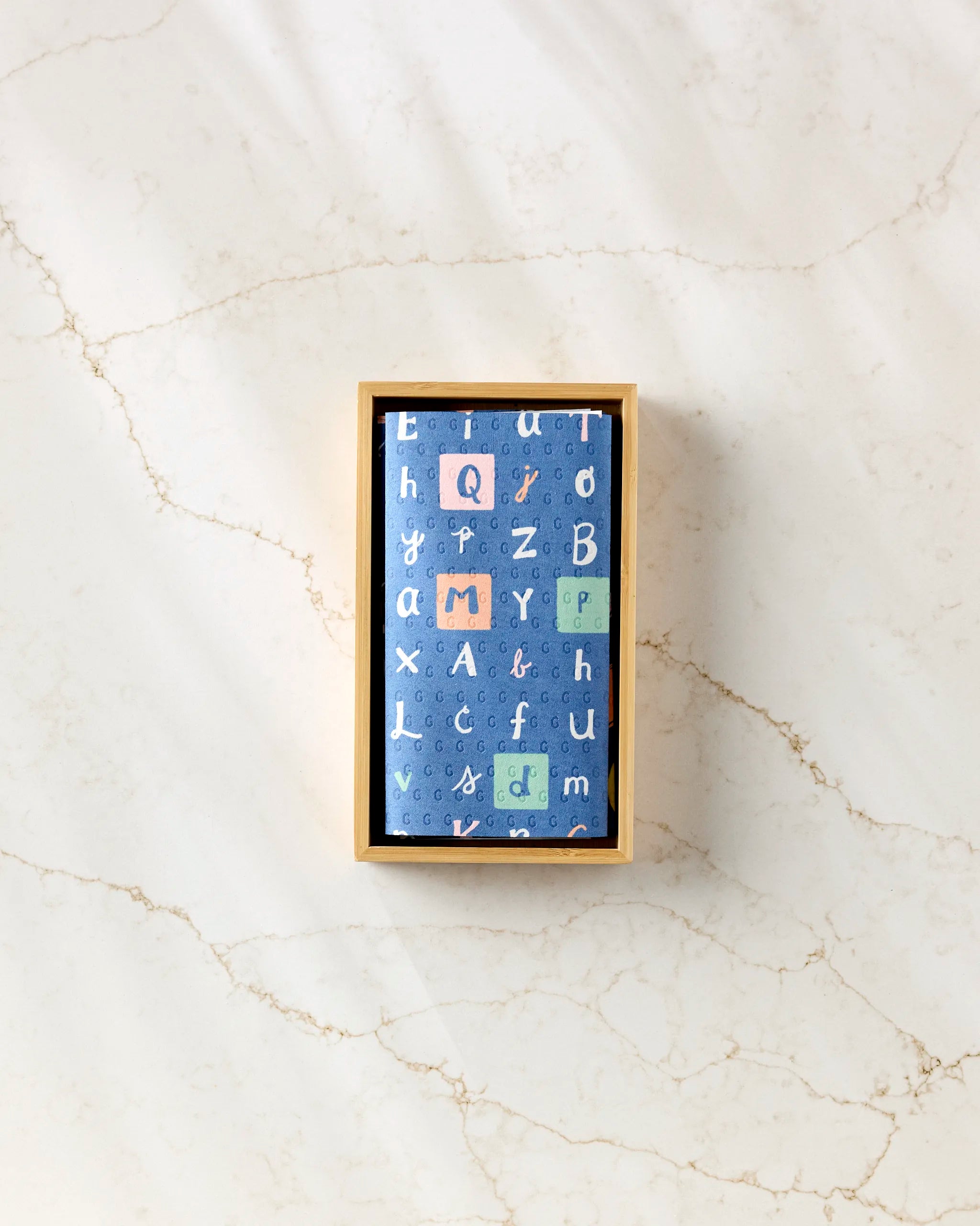 A rectangular "School Days" box by Amy MacCready, made from post-consumer recycled materials, has a blue lid with colorful alphabet letters in various sizes. The box sits on a light marble surface and is viewed from above.