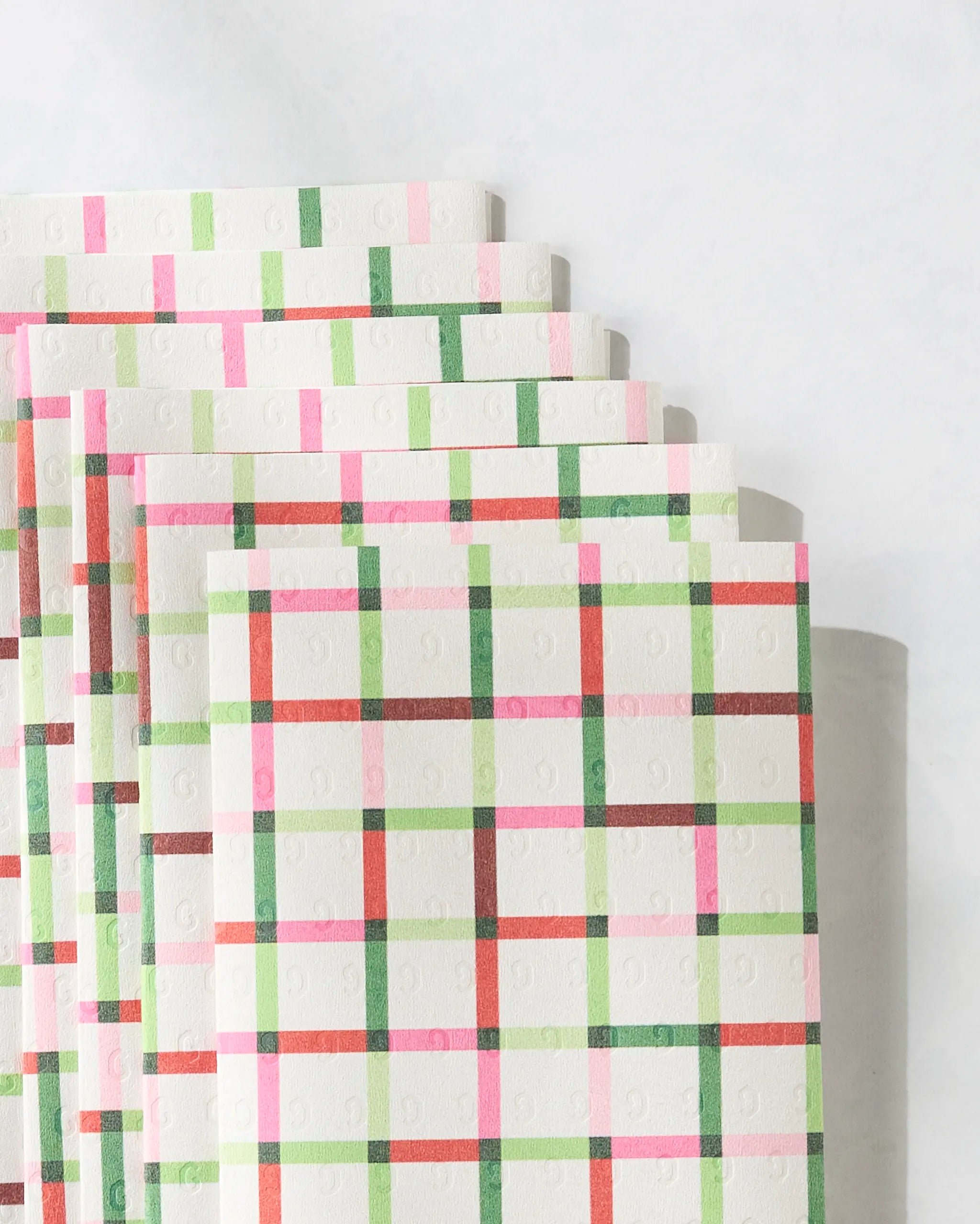 A close-up of Geometry’s Season of Joy eco-friendly paper towels, stacked in an overlapping fan shape, showcases a colorful red, green, and pink plaid pattern on a light background.