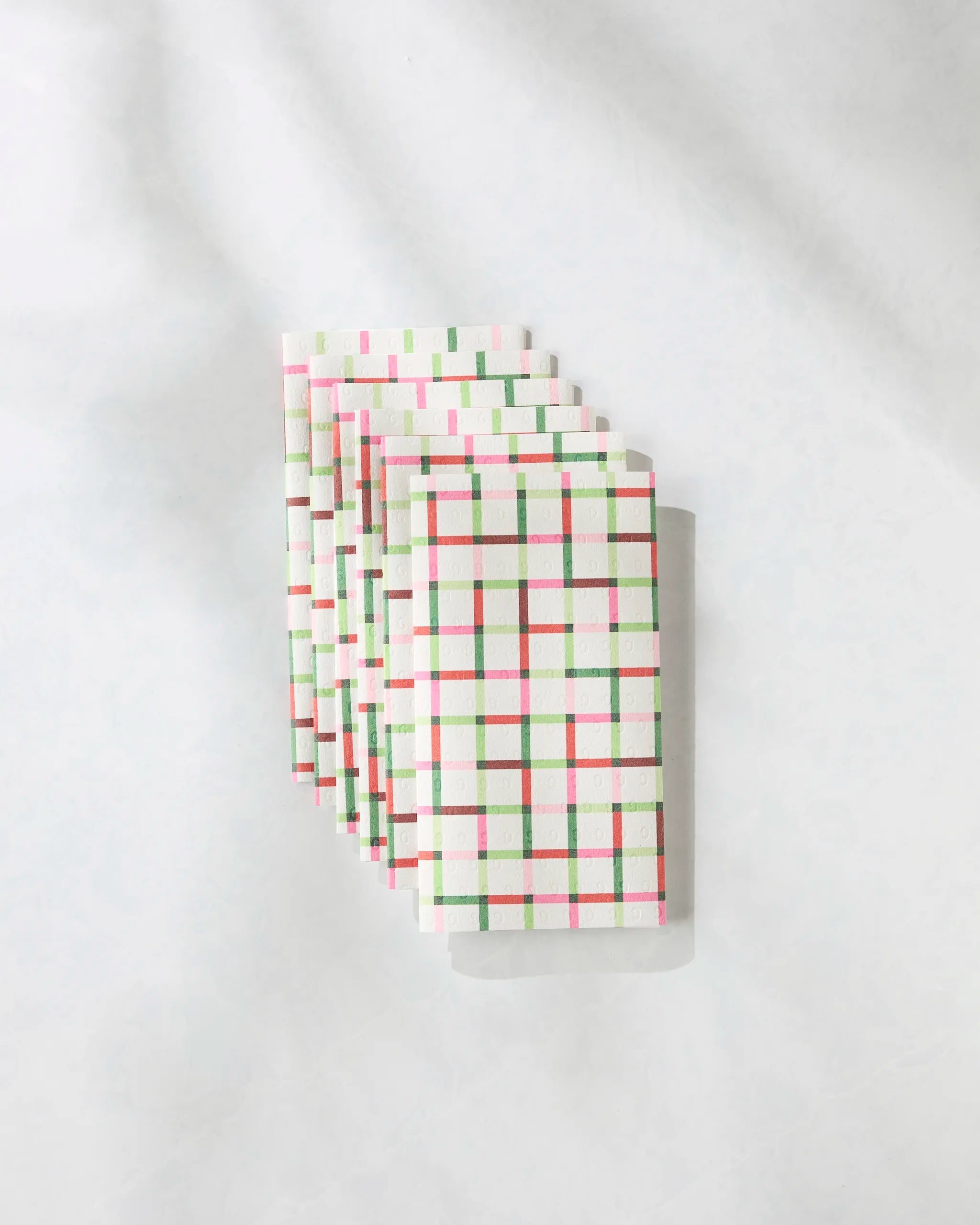 Seven folded "Season of Joy" reusable paper towels by Geometry, featuring red, green, and pink plaid, are stacked diagonally on a white surface with soft shadows.