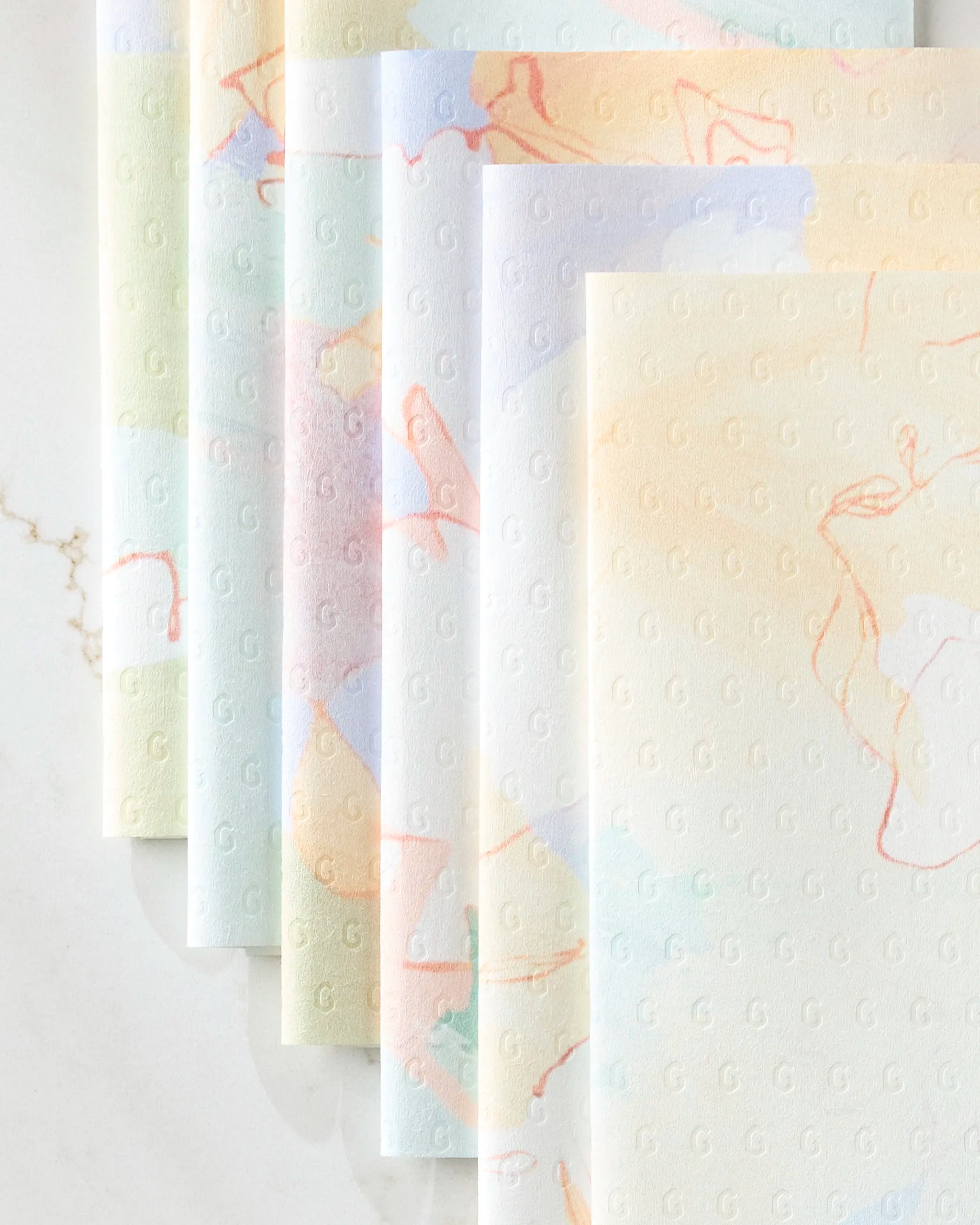 Secret Fort kitchen towels by Kristina Baker are eco-friendly and pastel-colored, with a subtle embossed “G” pattern and soft abstract swirls in light orange, blue, and green, neatly stacked on a marble surface.