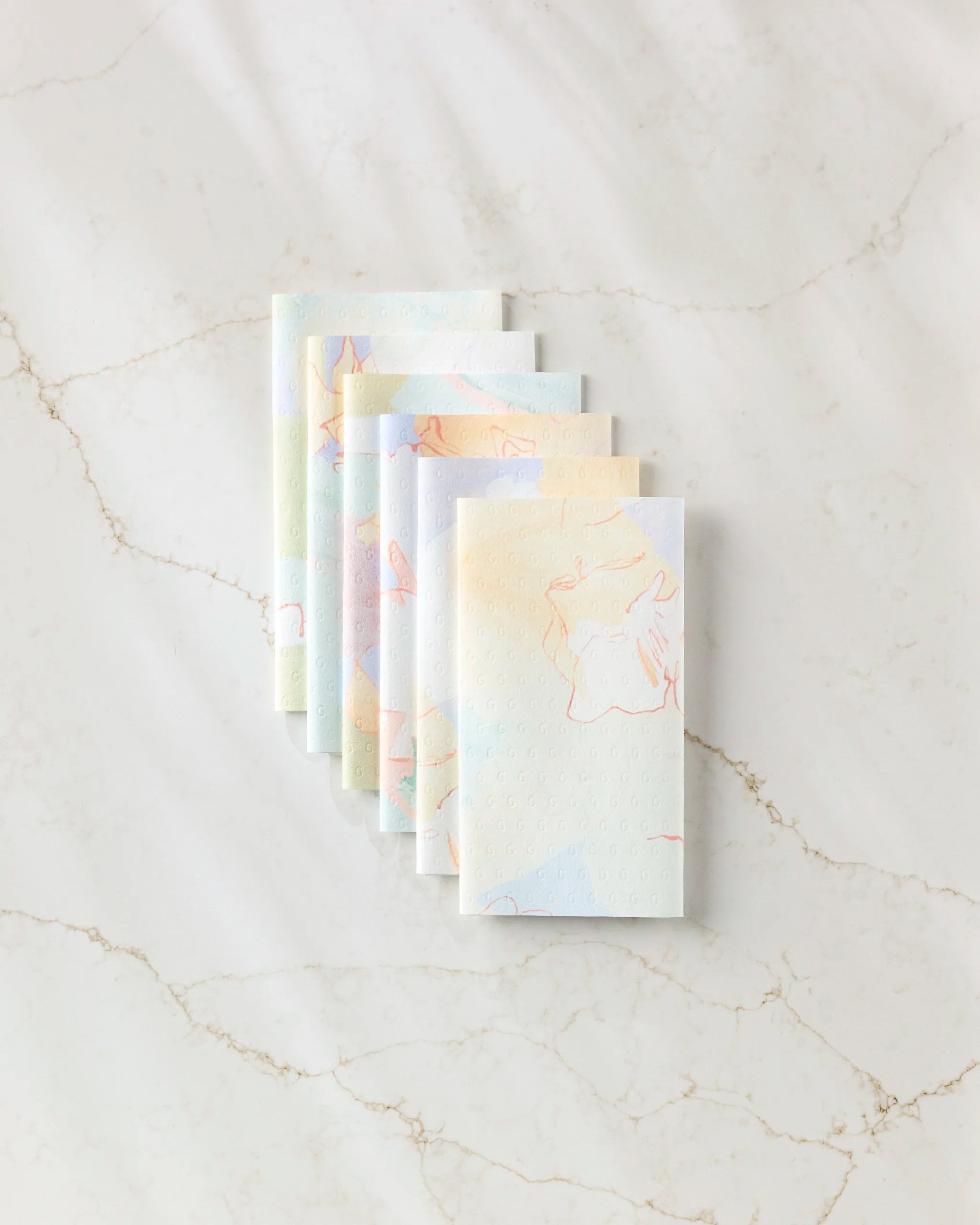Seven Secret Fort reusable paper towels by Kristina Baker, featuring a pastel abstract animal print, are neatly fanned out on a subtly veined white marble surface.