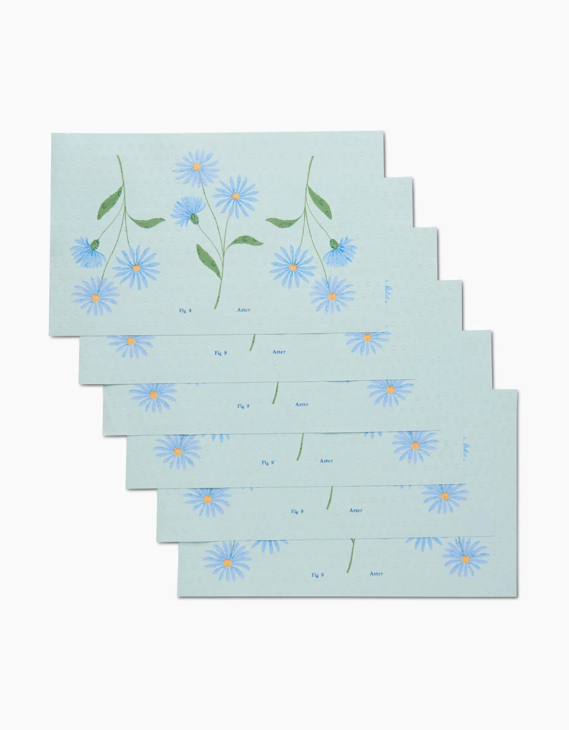 Six rectangular September Aster placemats by Linda Fox, each featuring blue aster flowers and green leaves, are fanned out for an eco-friendly table setting.