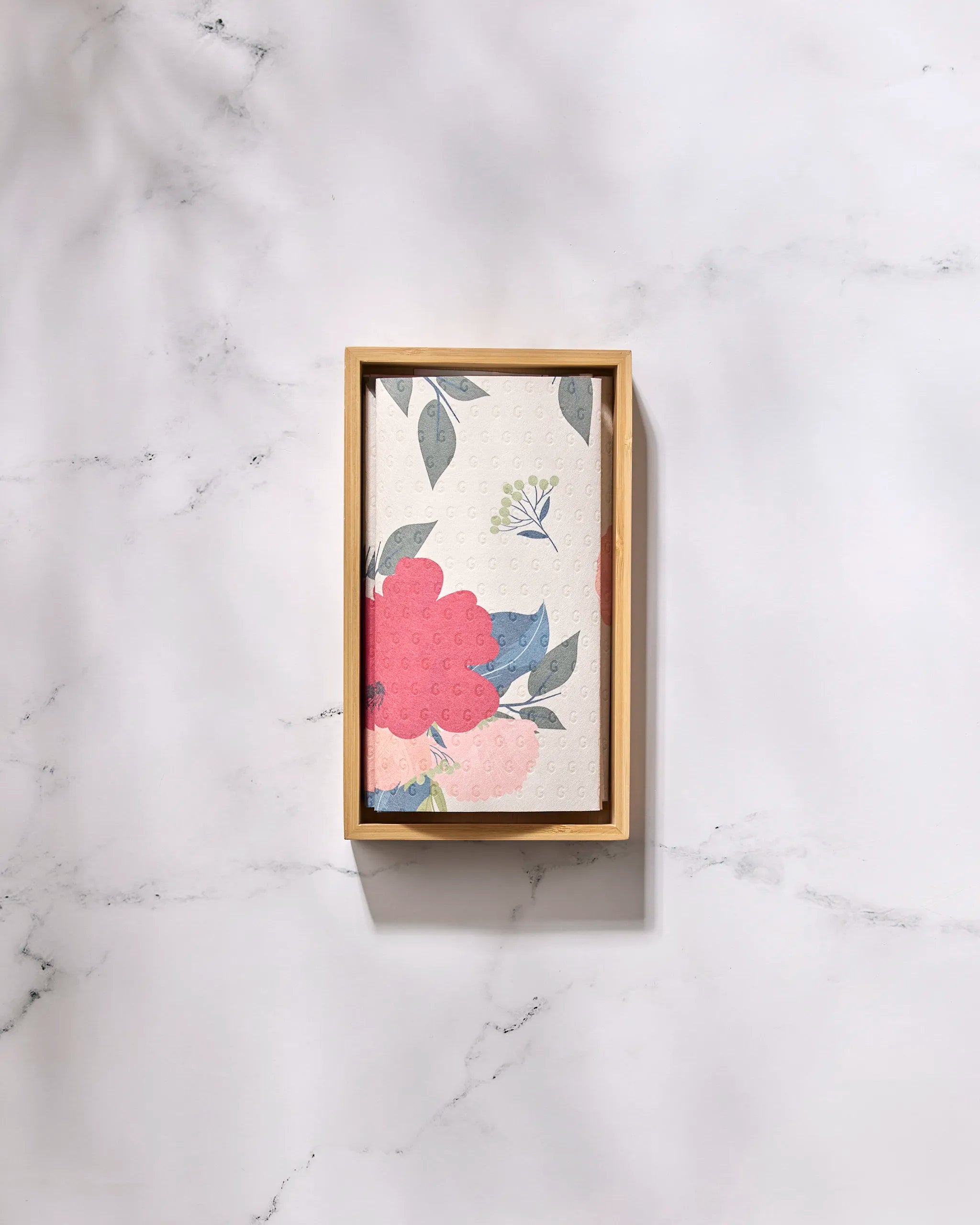 A folded "Showered With Love" napkin by GEOMETRY with pink and red floral designs, blue leaves, and colorful accents sits on a wooden tray—ideal as eco-friendly paper towels for a sustainable kitchen.