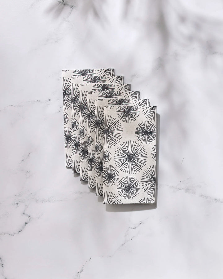 Five GEOMETRY Sky Party notebooks, featuring a black geometric sunburst pattern on white covers, are fanned out on a marble surface with soft shadows, reflecting the clean, modern style of eco-friendly kitchen towels.