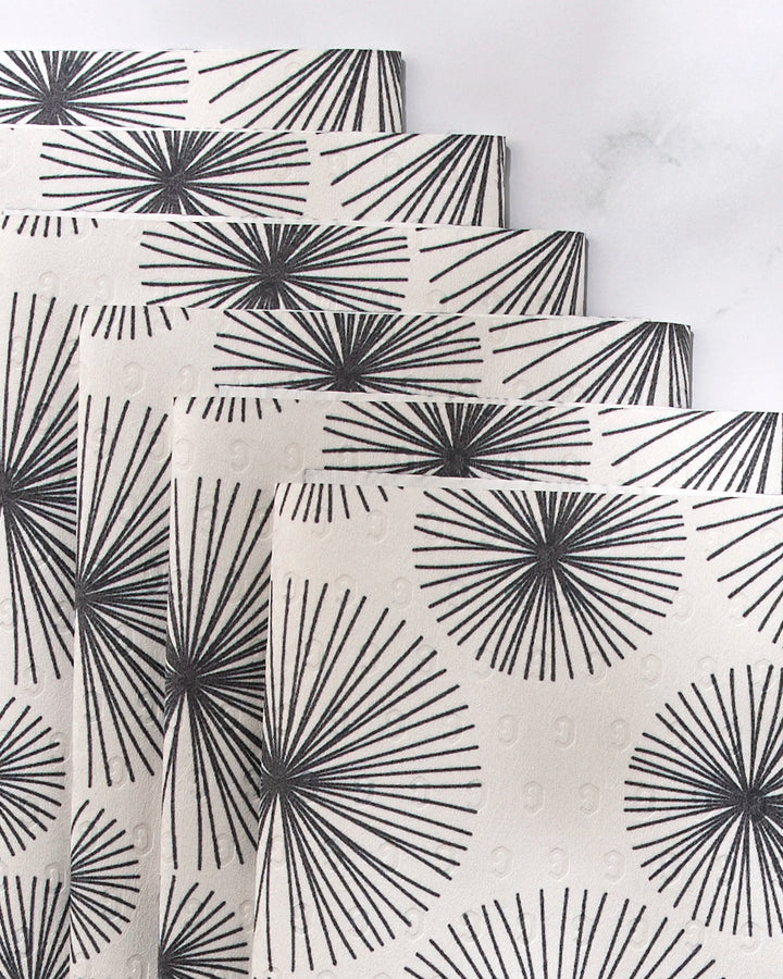 Five Sky Party reusable paper towels by GEOMETRY, featuring black, radiating line patterns on white and resembling abstract sunburst or dandelion shapes, are stacked in a staggered arrangement on a light, textured surface.