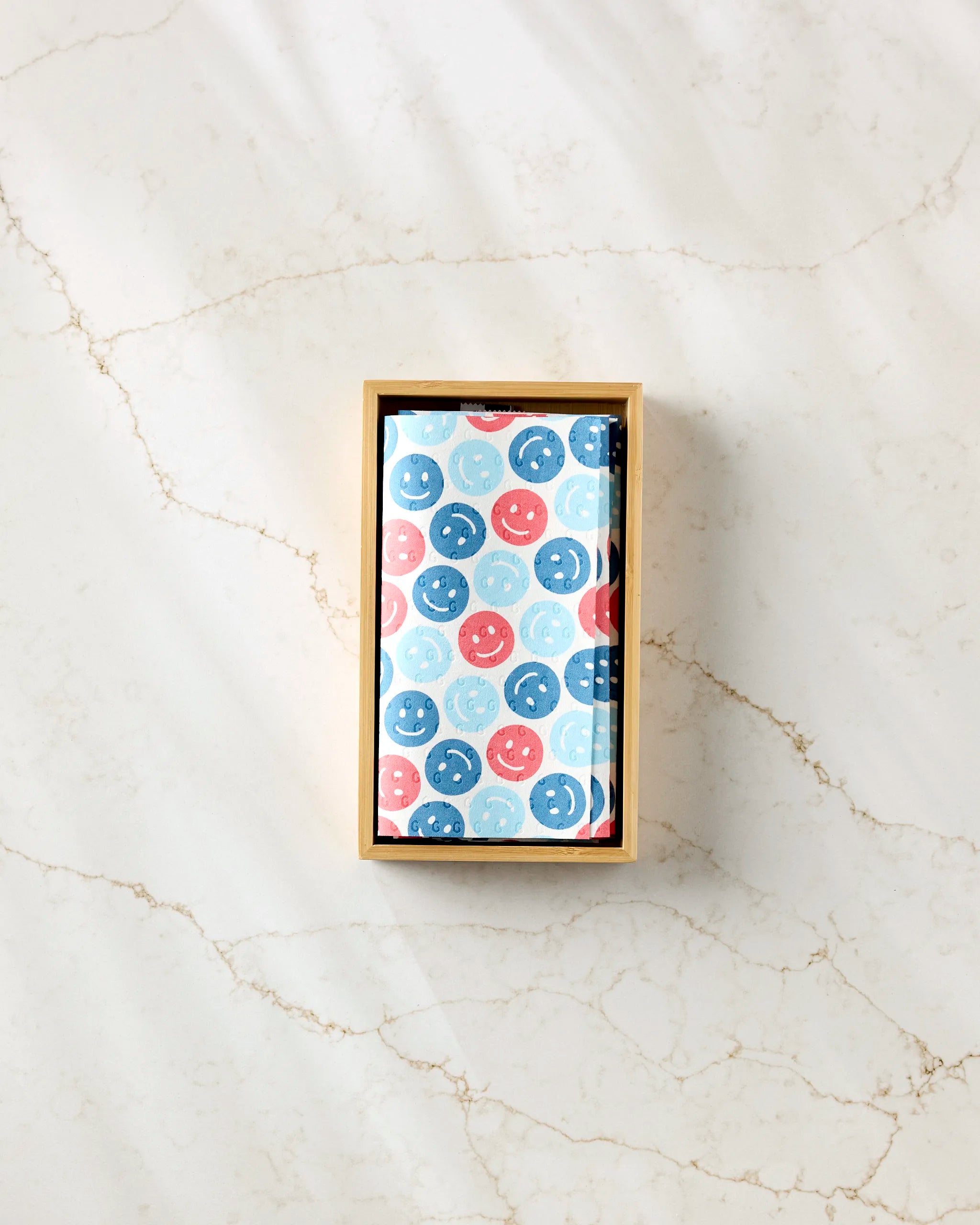 A rectangular box with a lid partly open reveals Hufton Studio's Smiley Face July Blue tissue paper—printed with blue, red, and light blue smiley faces—perfect for gifting eco-friendly kitchen towels on a light marble surface.