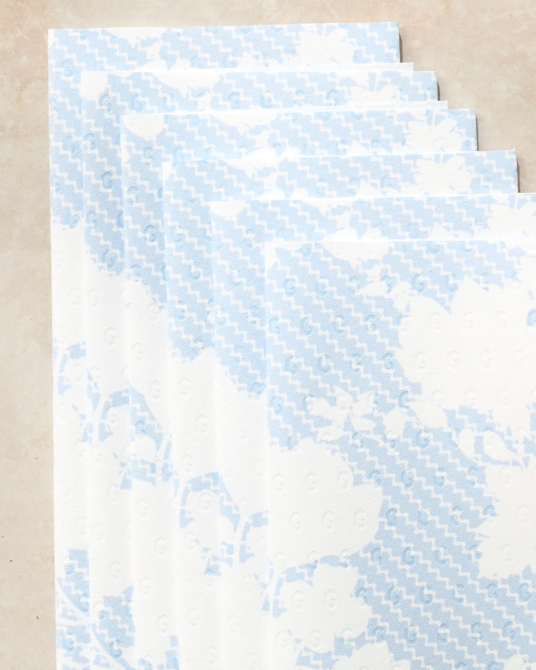 A close-up of several overlapping Geometry Soft Bloom Not Paper Towels, featuring a light blue floral and zigzag pattern, displayed on a beige surface.