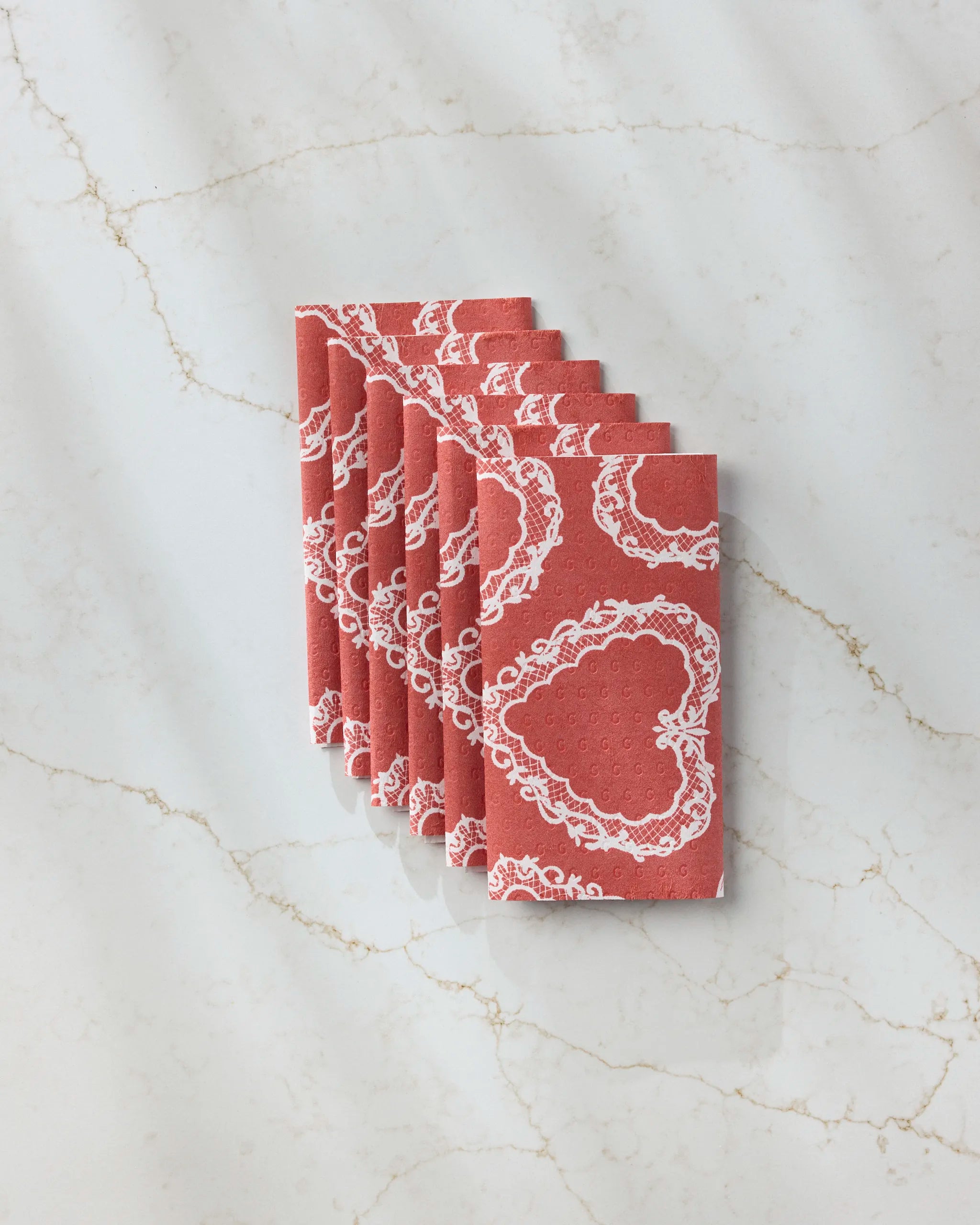 Eight folded Geometry Soft Heart Scarlet napkins, featuring white heart patterns and an eco-friendly paper towel design, are neatly arranged in a row on a white marble surface with light brown veining.