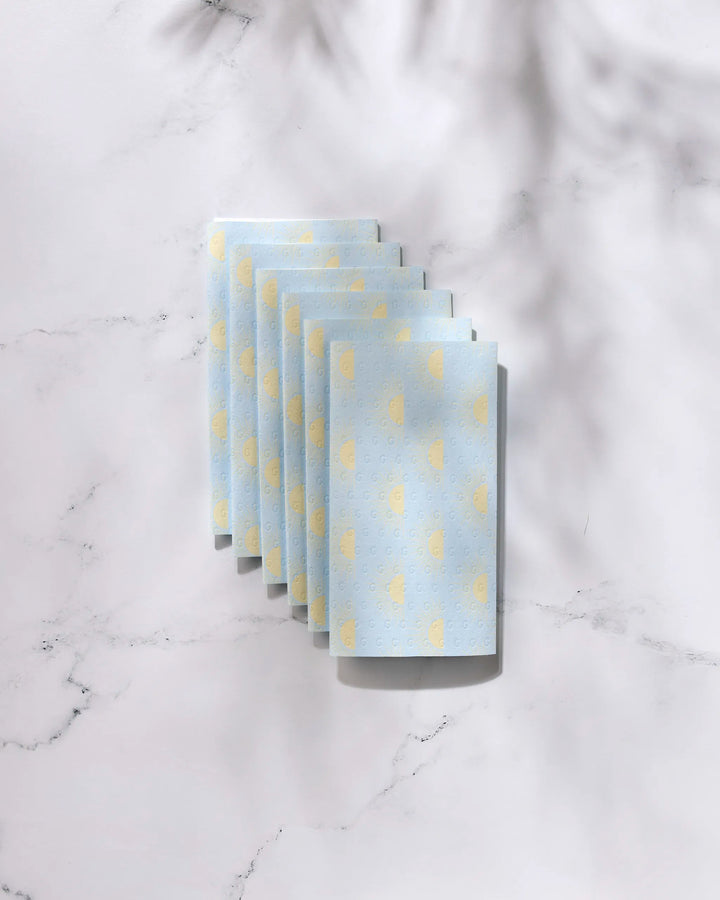 Six folded GEOMETRY Soft Summer Sun blue reusable paper towels with yellow polka dots are arranged in a neat, overlapping row on a white marble surface with soft shadows.