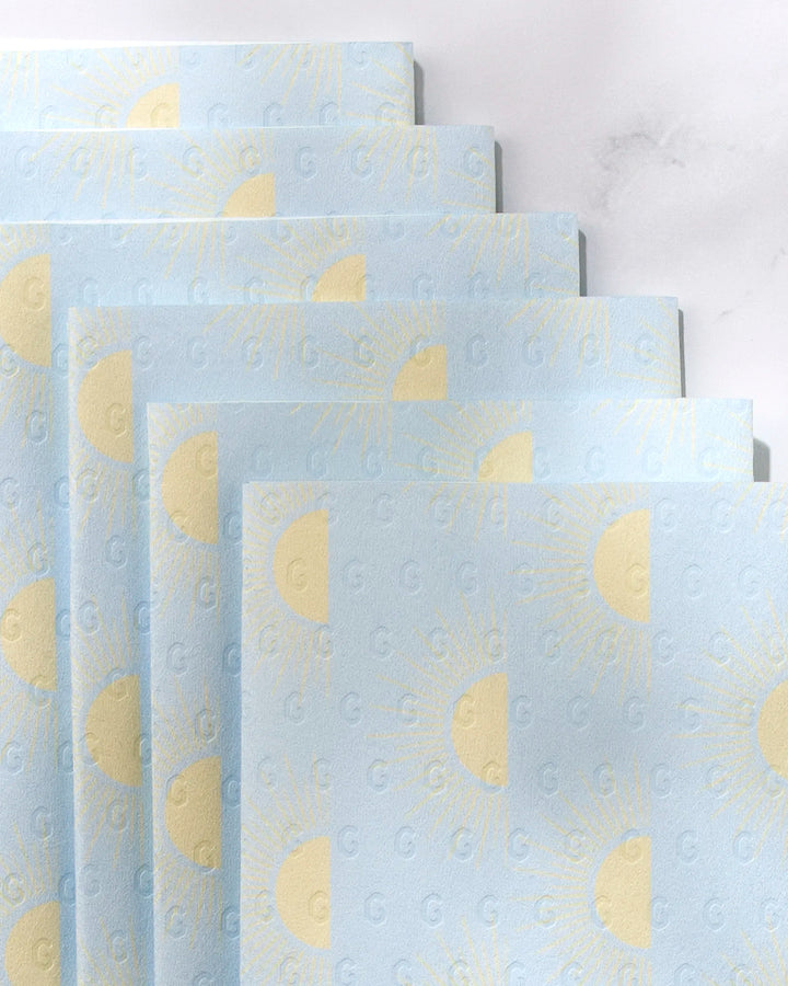 Several sheets of GEOMETRY's Soft Summer Sun eco-friendly, light blue paper towels—decorated with yellow suns and a subtle “G” emboss—are arranged in a staggered, overlapping layout on a light background.