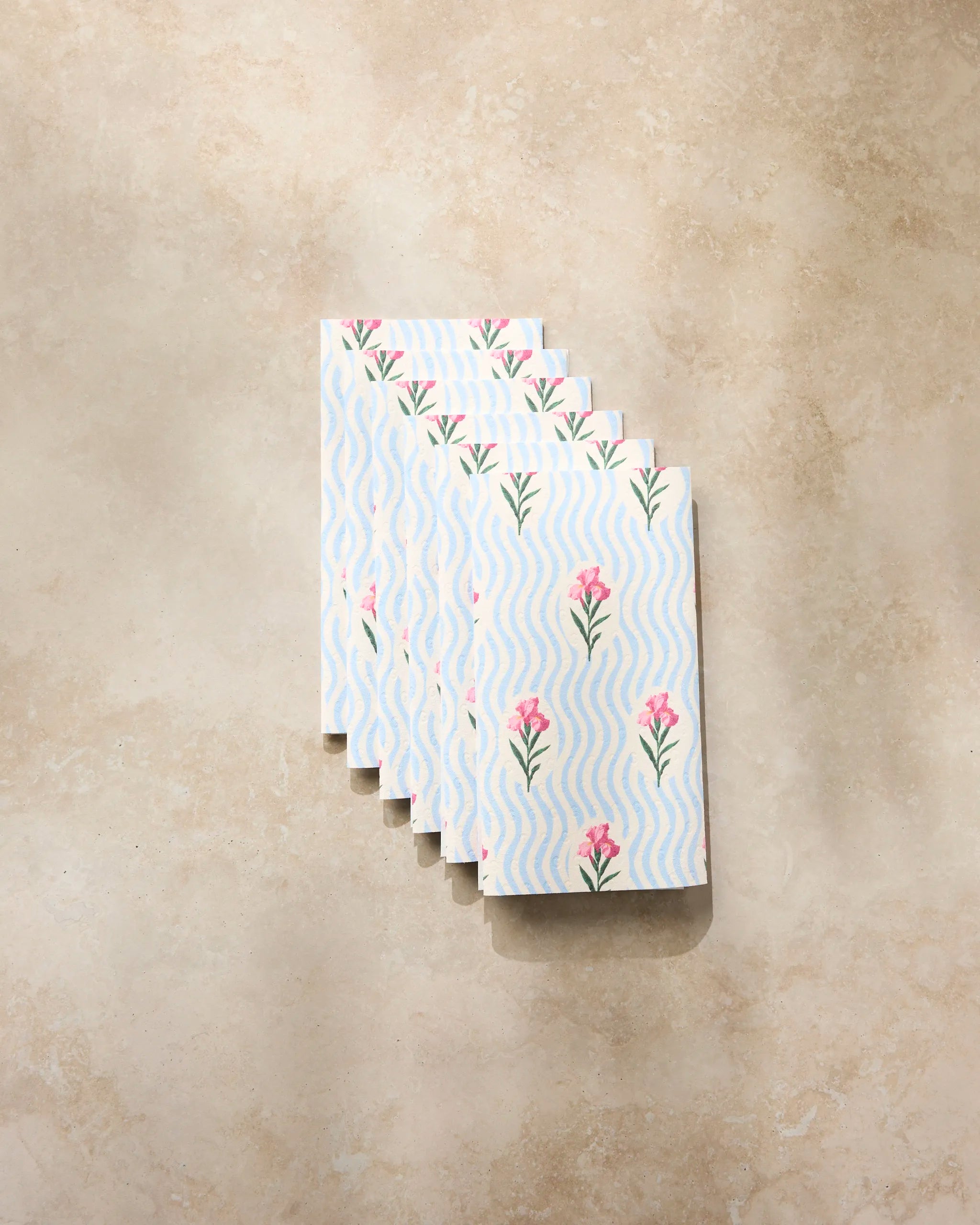 Five "Spatial Bloom" small notebooks by Geometry, featuring a wavy blue-and-white design with pink flowers, are fanned out on a beige textured surface, evoking the look of eco-friendly kitchen towels.