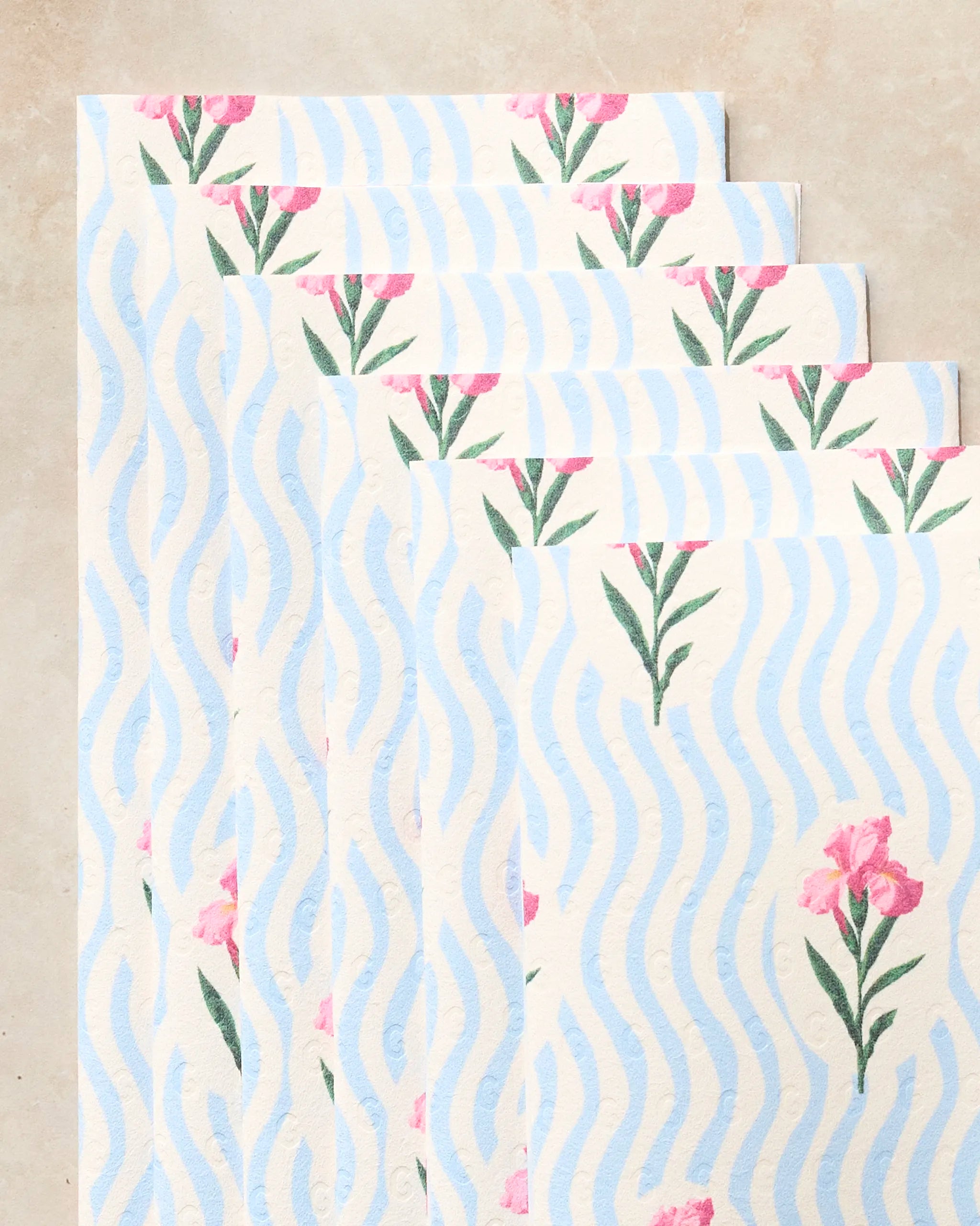 Five sheets of Geometry's Spatial Bloom kitchen towels, featuring a wavy blue stripe pattern with small pink flowers and green stems, are fanned out on a light beige surface.
