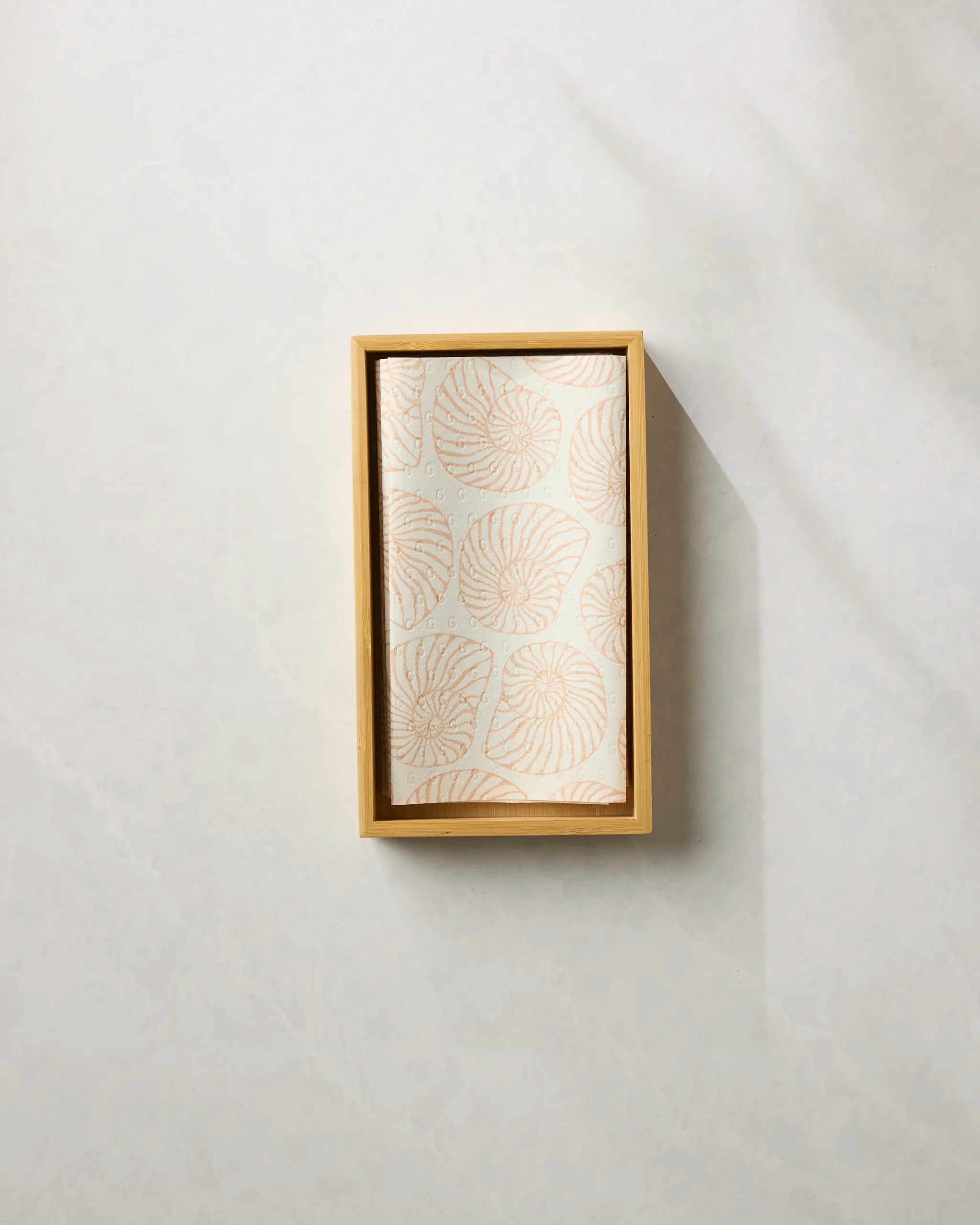 The Spira reusable paper towel by Geometry, featuring a light beige leaf pattern, is neatly folded inside a rectangular wooden tray on a light-colored textured surface.