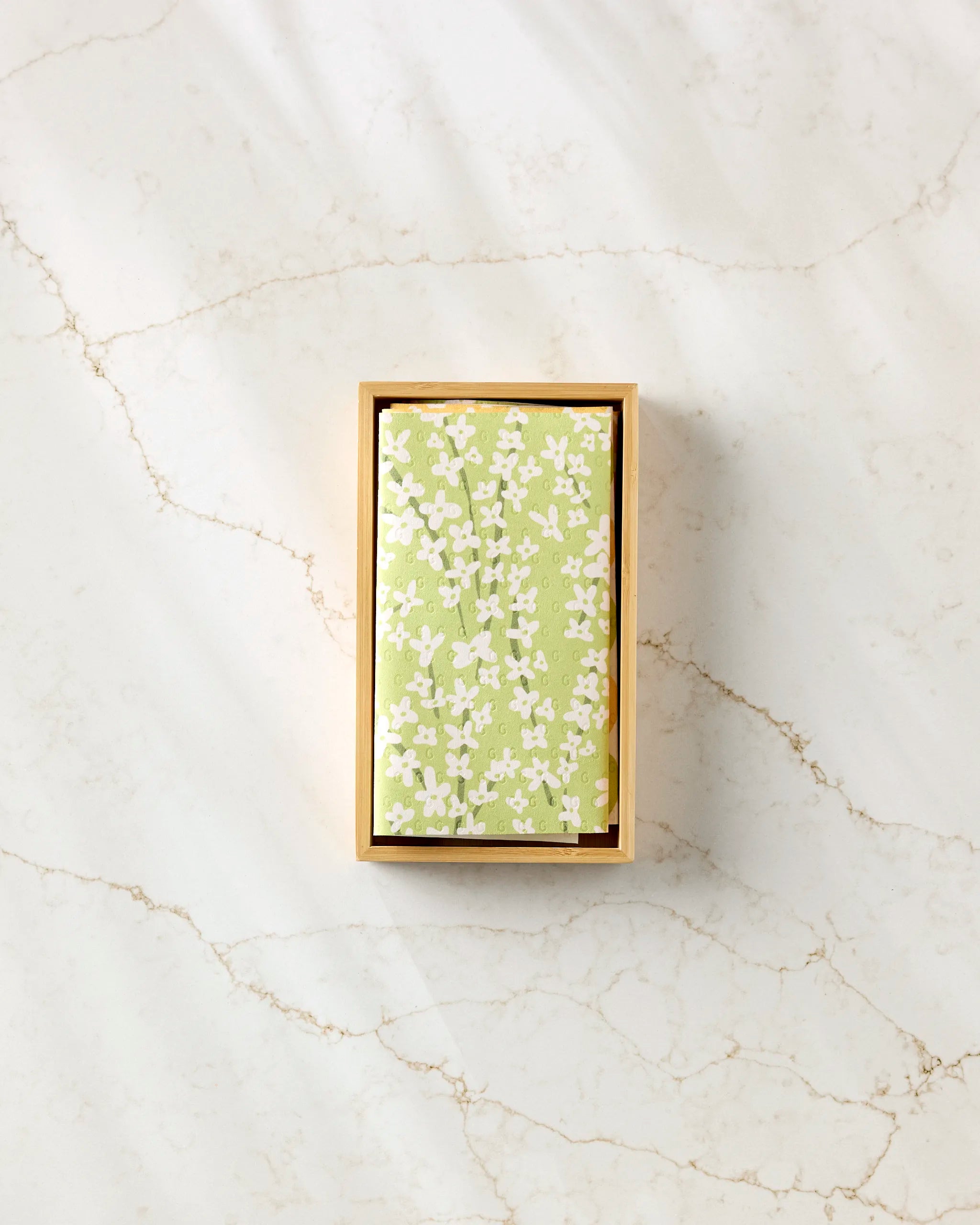 The "Spring Bounty" box by Kayla Ann, featuring a green lid with a white floral pattern, is designed for eco-friendly kitchen towels and is displayed on a light marble surface with tan veins.