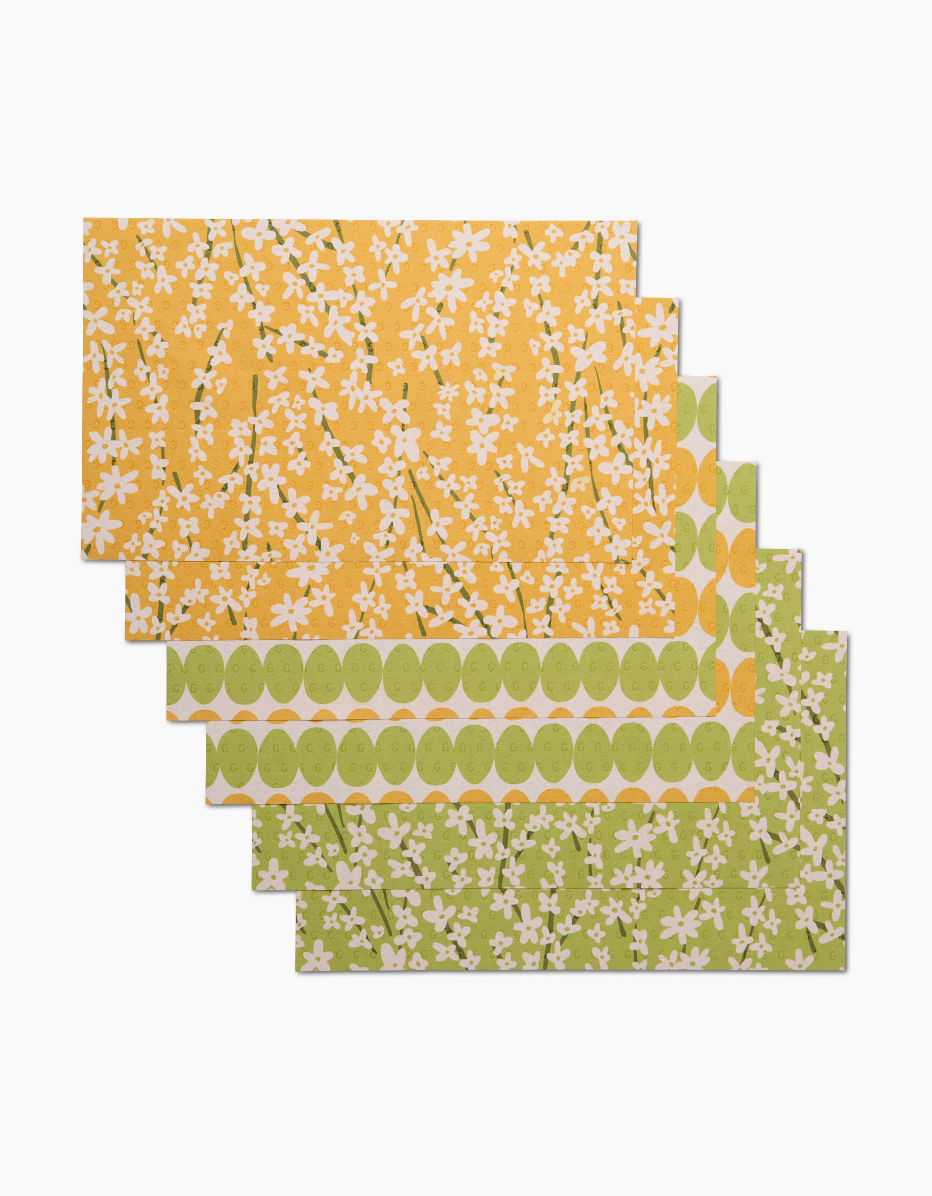 Four Kayla Ann Spring Bounty rectangular placemats, featuring floral and geometric designs in yellow, green, and white, are stacked with the top two in yellow and the bottom two in green. They resemble reusable kitchen towels.