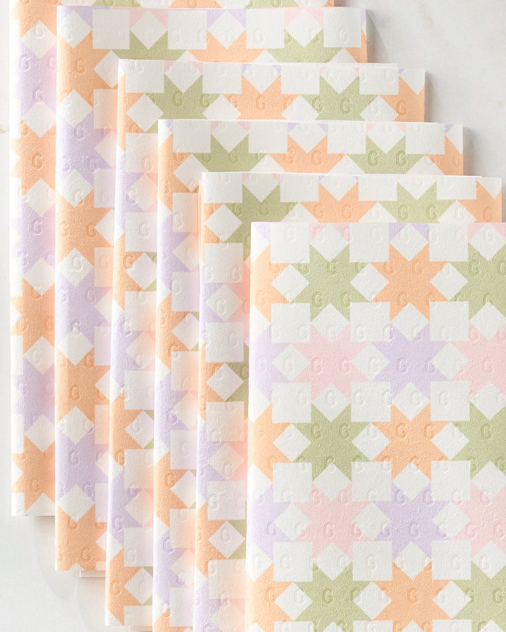 Five folded Kayla Ann Spring Quilt kitchen towels in pastel peach, lavender, green, and white with a geometric diamond and arrow pattern are arranged in an overlapping diagonal stack on a light surface.