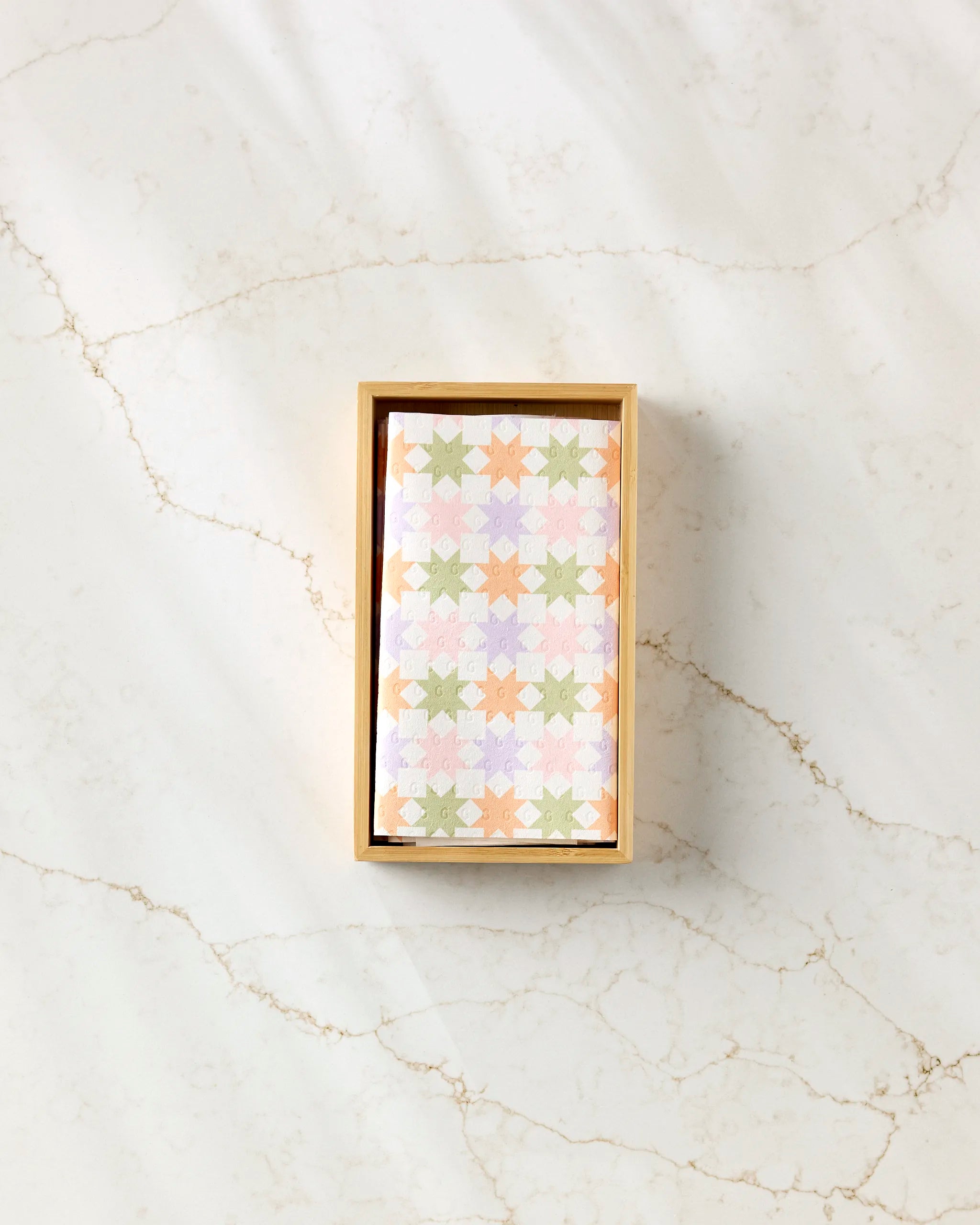 The Kayla Ann Spring Quilt, featuring a pastel star pattern in green, orange, yellow, pink, and lavender, rests folded in a wooden box—ideal as eco-friendly kitchen towels or reusable paper towels for sustainable living.
