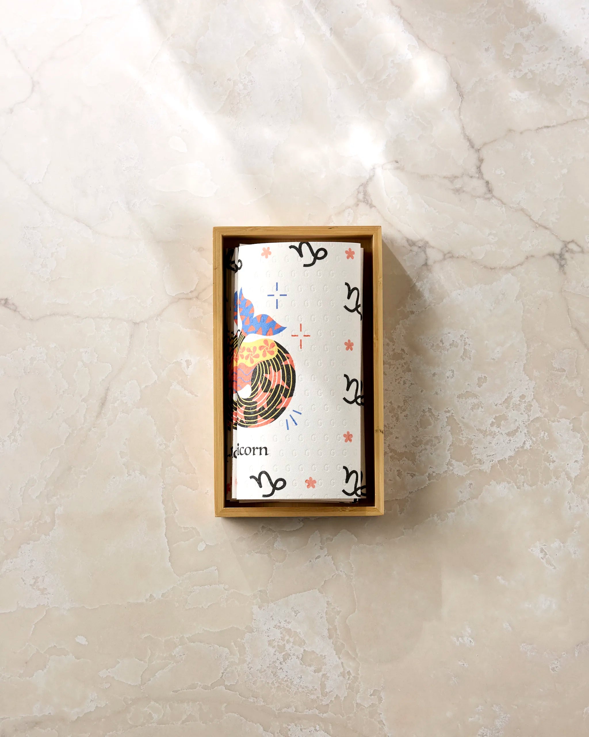 A rectangular box labeled "Steadfast Capricorn" by Heather Zhou sits on marble, containing a card with abstract patterns and zodiac symbols. Sunlight casts soft shadows near neatly stacked eco-friendly paper towels.