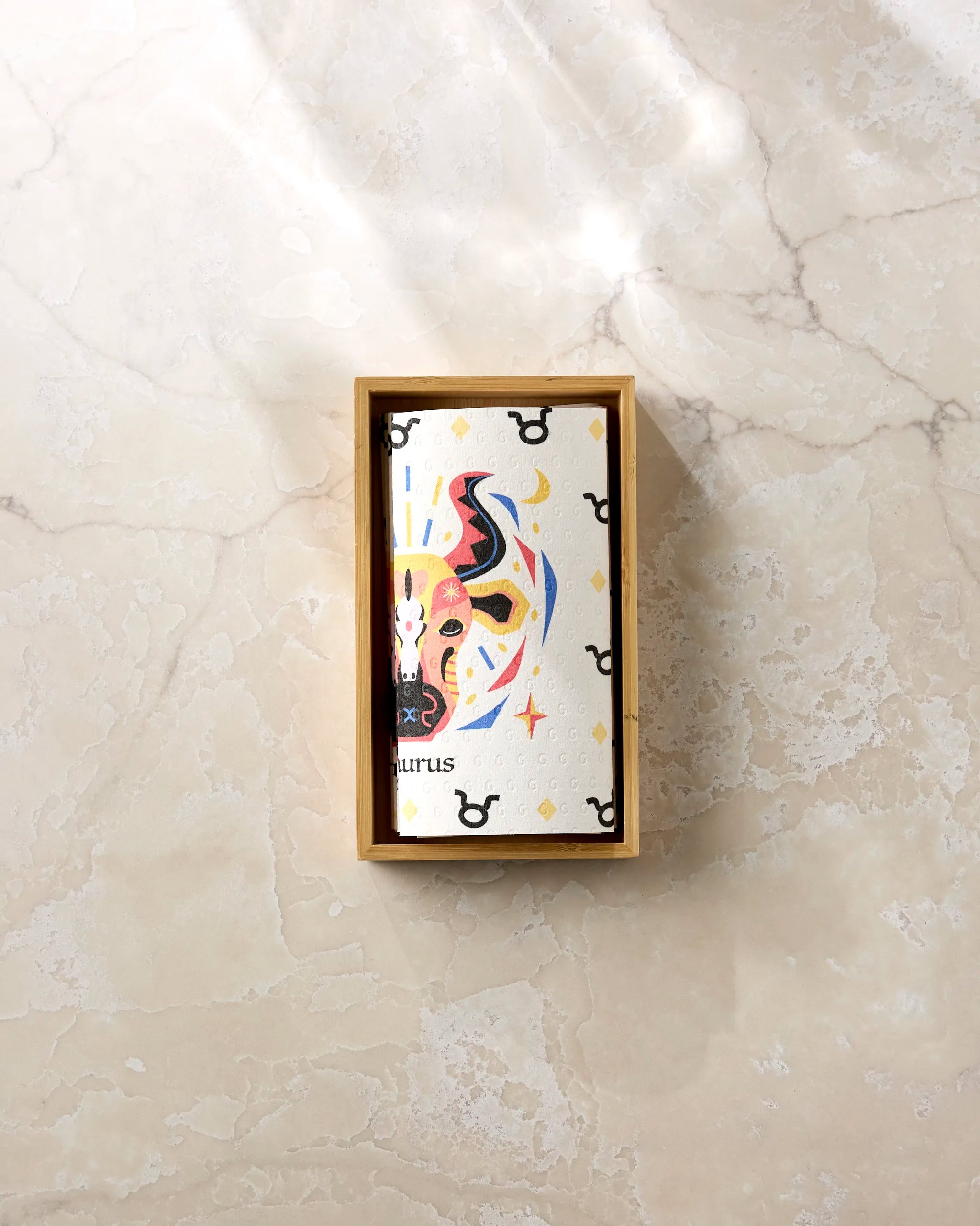 A vibrant Steady Taurus card by Heather Zhou, featuring a stylized bull and astrological symbols, rests in a wooden box on a marbled surface next to eco-friendly kitchen towels. The word 'Taurus' appears on the card.