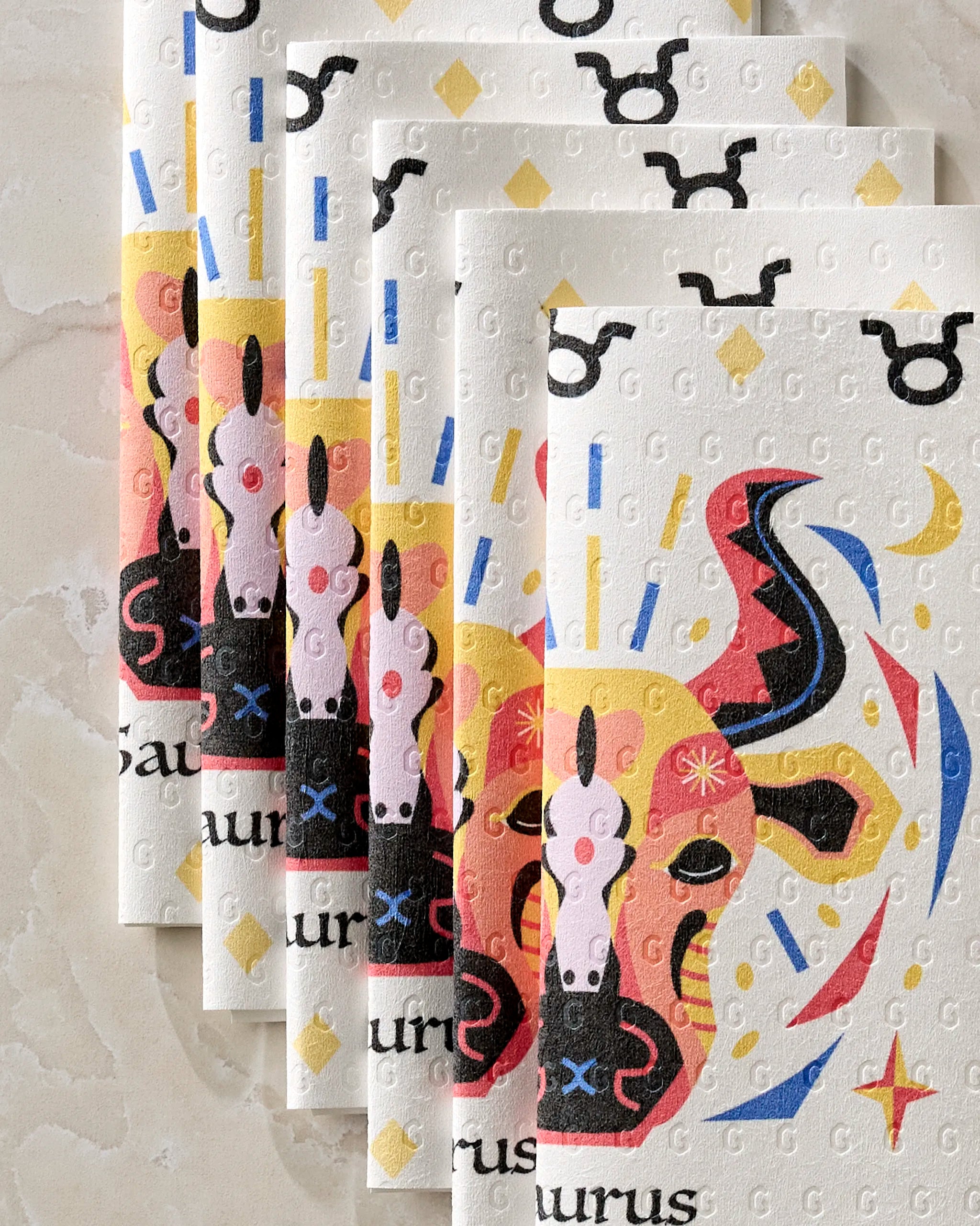 Five Steady Taurus reusable paper towels by Heather Zhou are stacked on a light marble surface, each displaying a colorful Taurus bull with abstract patterns and symbols in red, yellow, black, and blue.