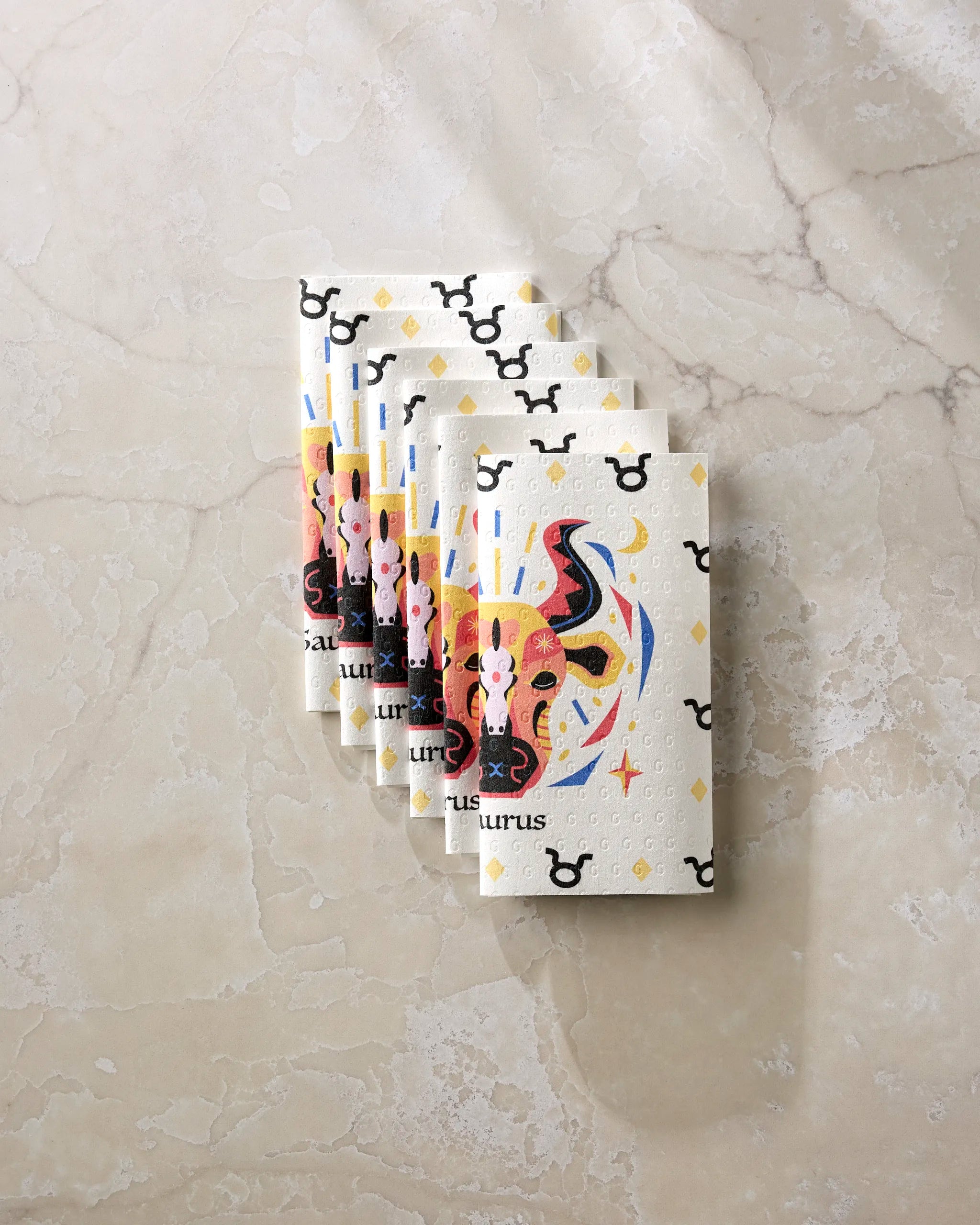 Six Steady Taurus cards by Heather Zhou display vibrant bull art and Taurus zodiac symbols on a marble surface. Water droplets on the cards evoke the freshness of eco-friendly kitchen towels after sustainable cleaning.