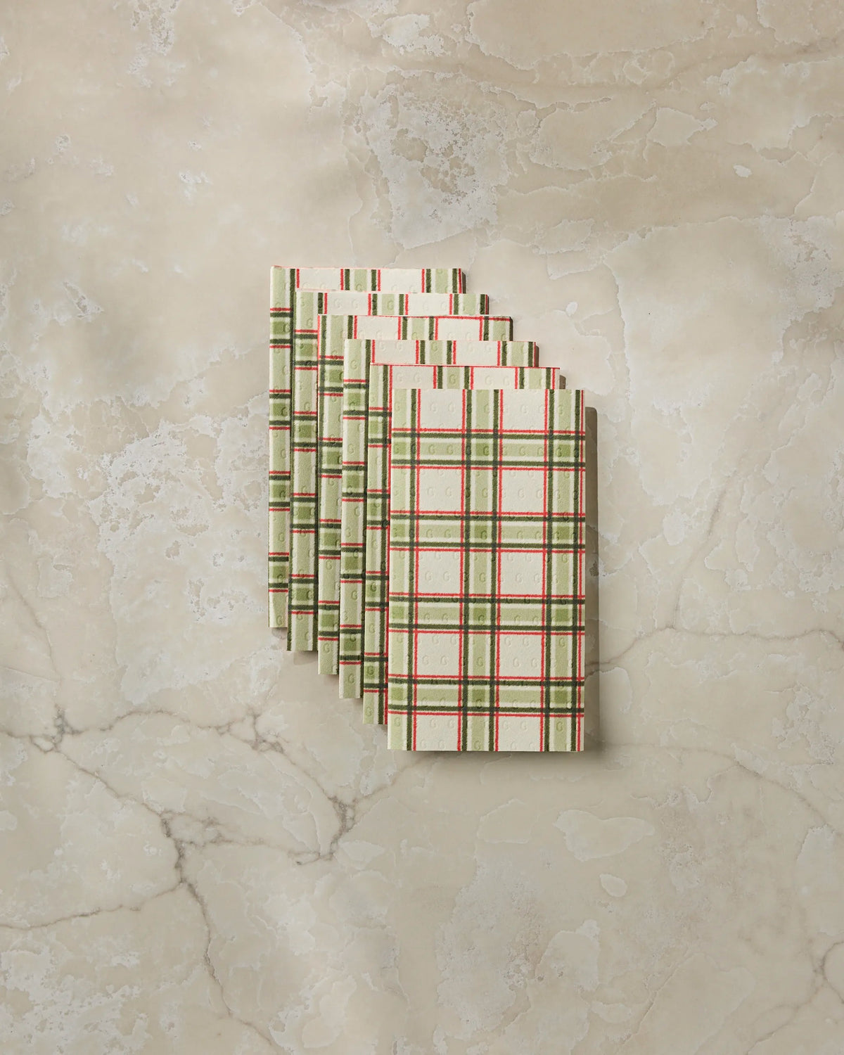 Six Stocking Plaid chocolate bars by Geometry, featuring cream, green, and red plaid patterns inspired by the classic designs of eco-friendly kitchen towels, are stacked and fanned out on a beige marble surface.