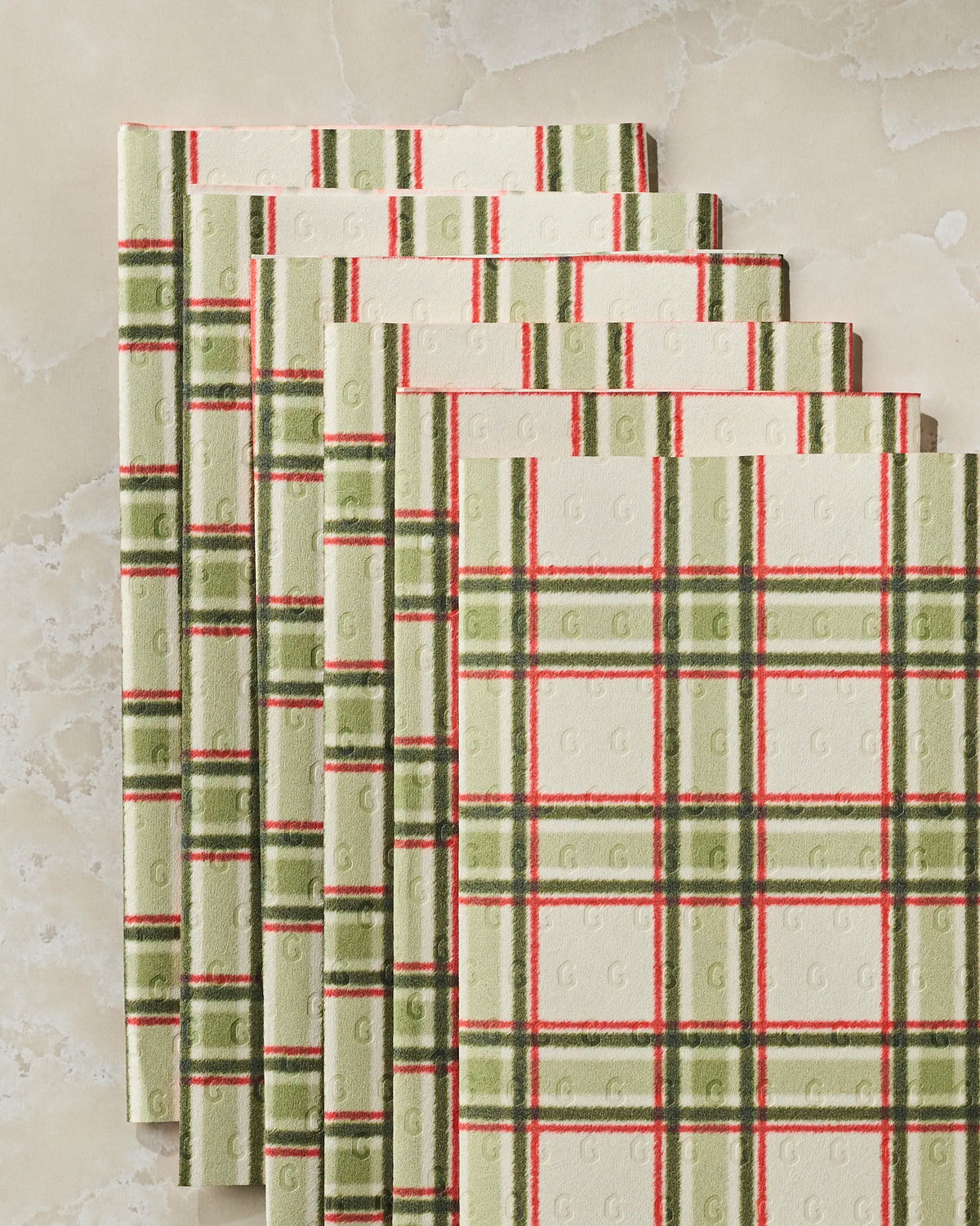 Five Stocking Plaid napkins by Geometry, featuring green, red, and cream plaid with a subtle G pattern, are stacked diagonally on a light marbled surface. These versatile napkins double as eco-friendly kitchen towels, adding a sustainable touch to your dining or cleaning routine.