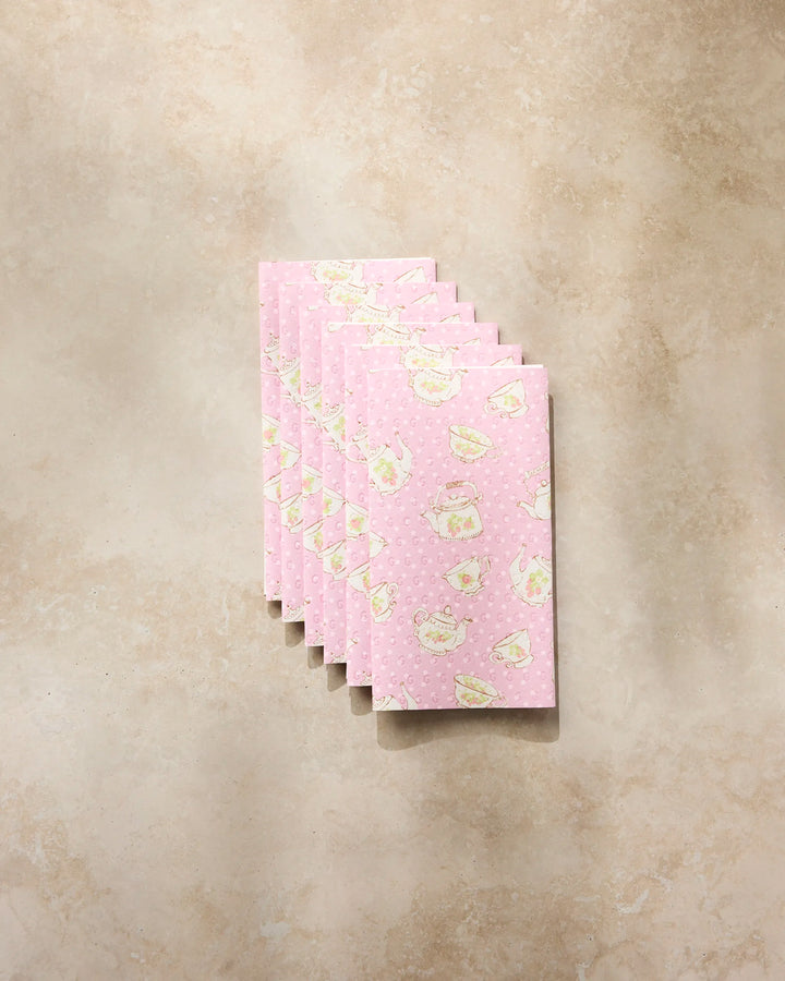 Seven pink reusable paper towels from Geometry, featuring the "Strawberry Tea" design with white teacup and teapot motifs, are neatly fanned out on a beige, textured surface.