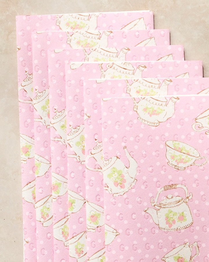 Five pastel pink “Strawberry Tea” napkins by Geometry, not paper towels, display teapots, cups, and saucers with floral designs in a fanned-out stack on a light surface.