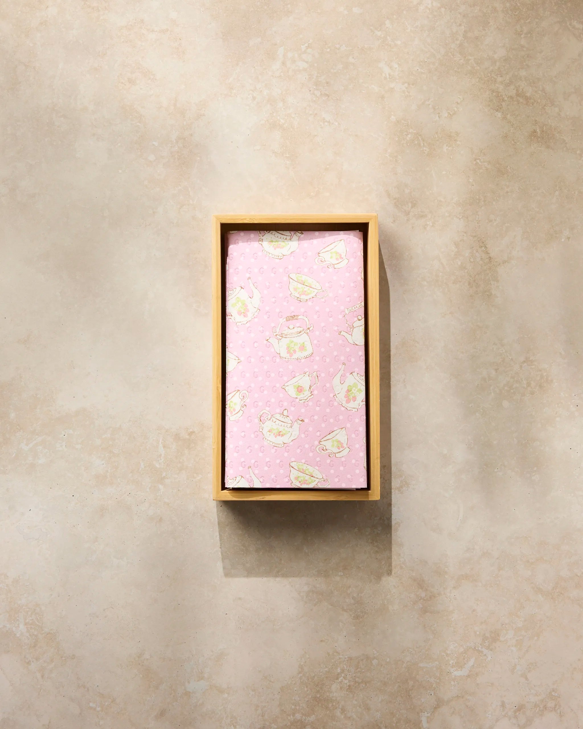 A rectangular wooden box holds a folded pink Strawberry Tea towel by Geometry, featuring white cartoon cats and yellow stars, displayed against a light, textured background.