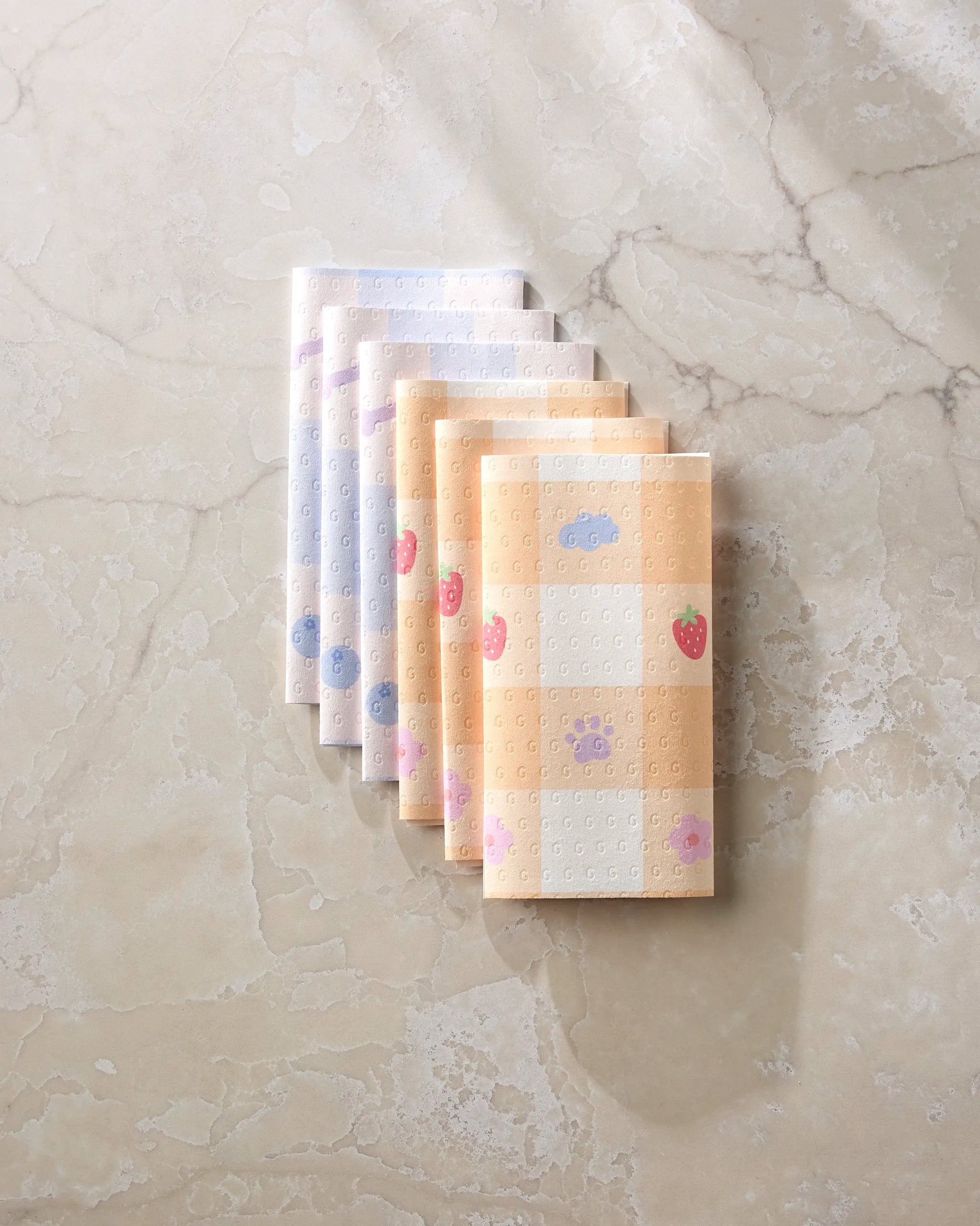 Seven folded Summer Picnic Dreams reusable towels by Letters By Shells, featuring pastel blue, purple, and yellow checkered patterns with fruit and animal designs, are neatly arranged as an eco-friendly kitchen option.