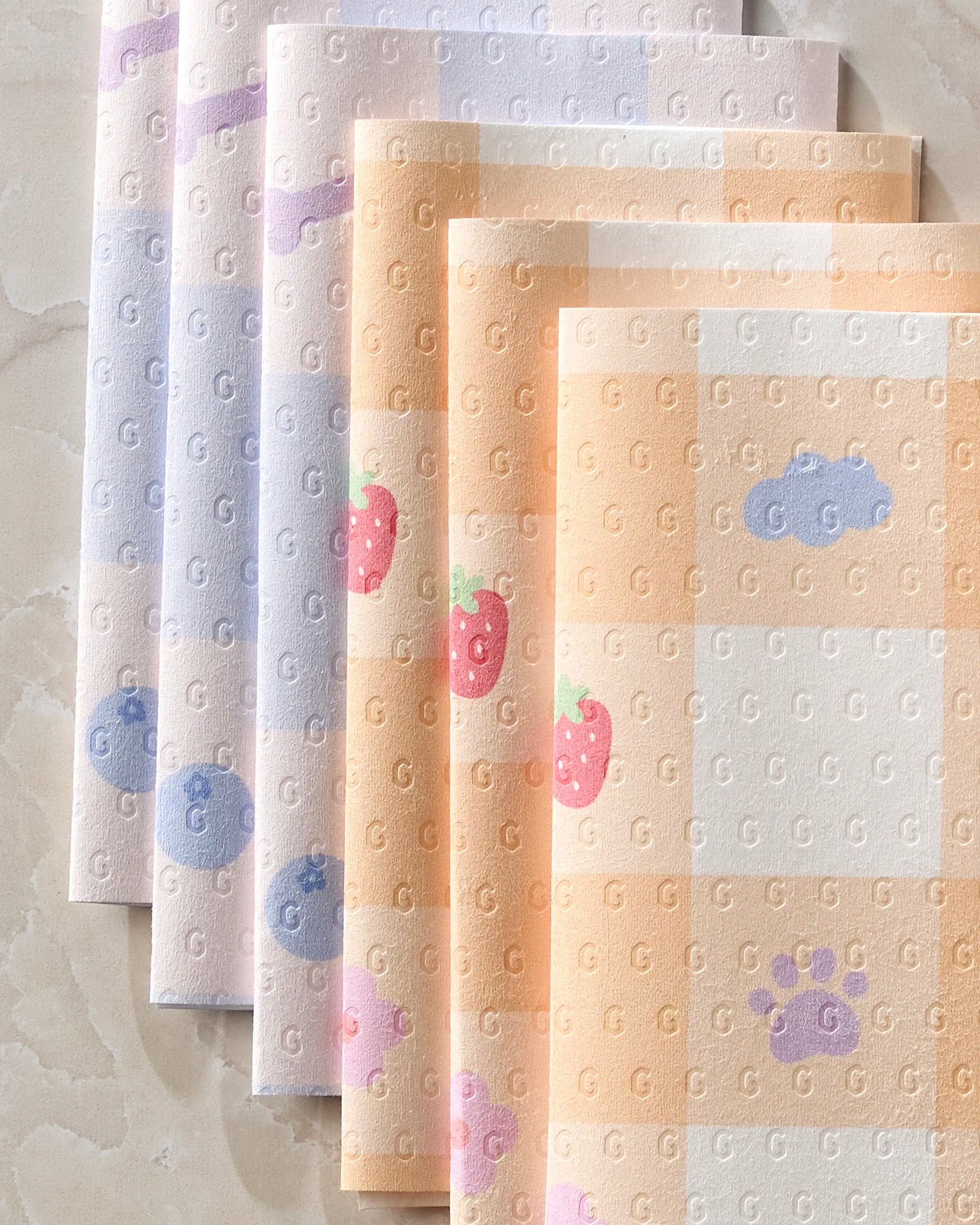 Summer Picnic Dreams by Letters By Shells features five eco-friendly kitchen towels with pastel checkered patterns, adorned with clouds, strawberries, and paw prints in soft orange, purple, and pink. Sheets are arranged in an overlapping display.
