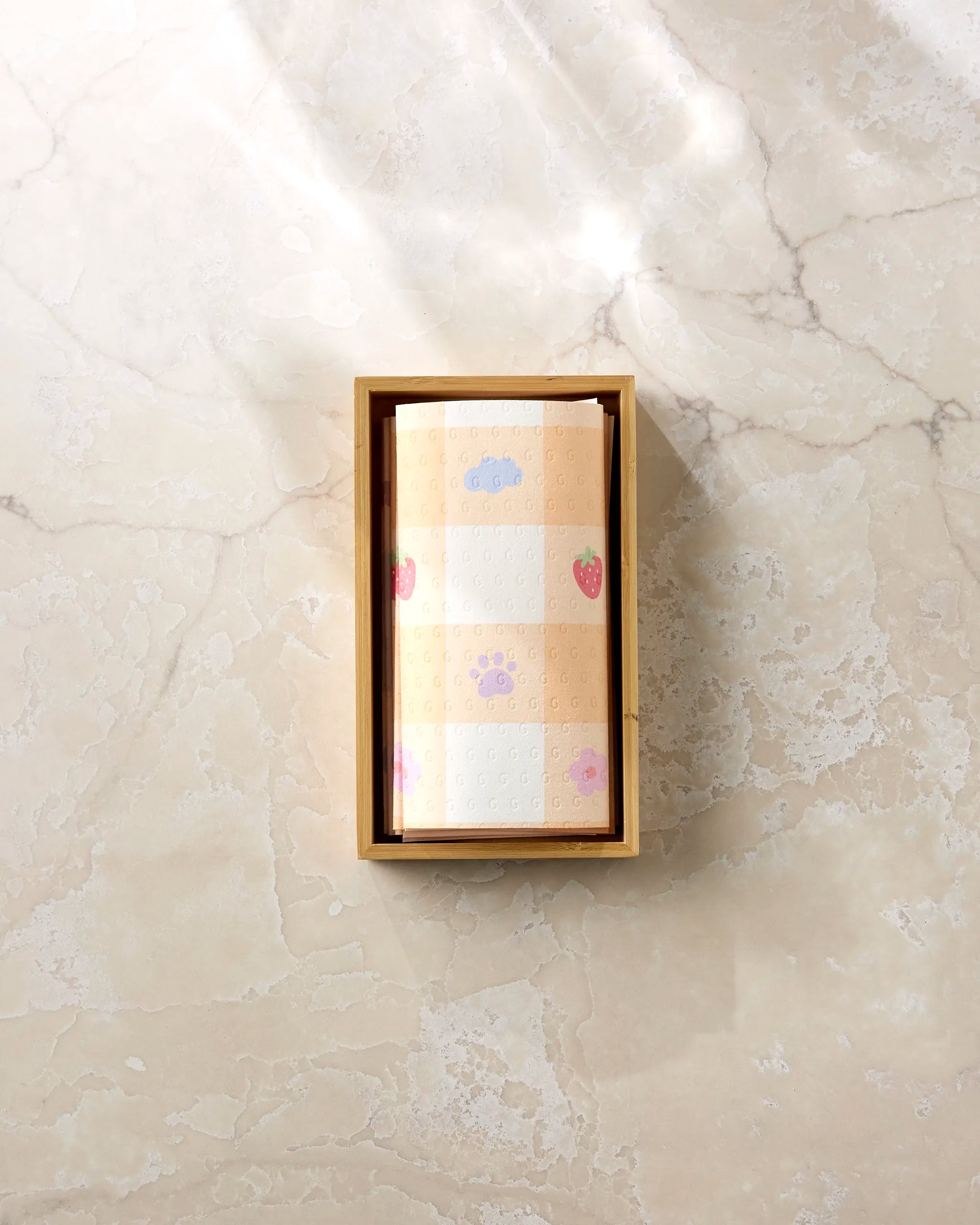 A rectangular wooden box holds folded beige Summer Picnic Dreams kitchen towels by Letters By Shells, featuring checkered patterns and small prints of strawberries, clouds, and paw prints, displayed on a light marble surface.