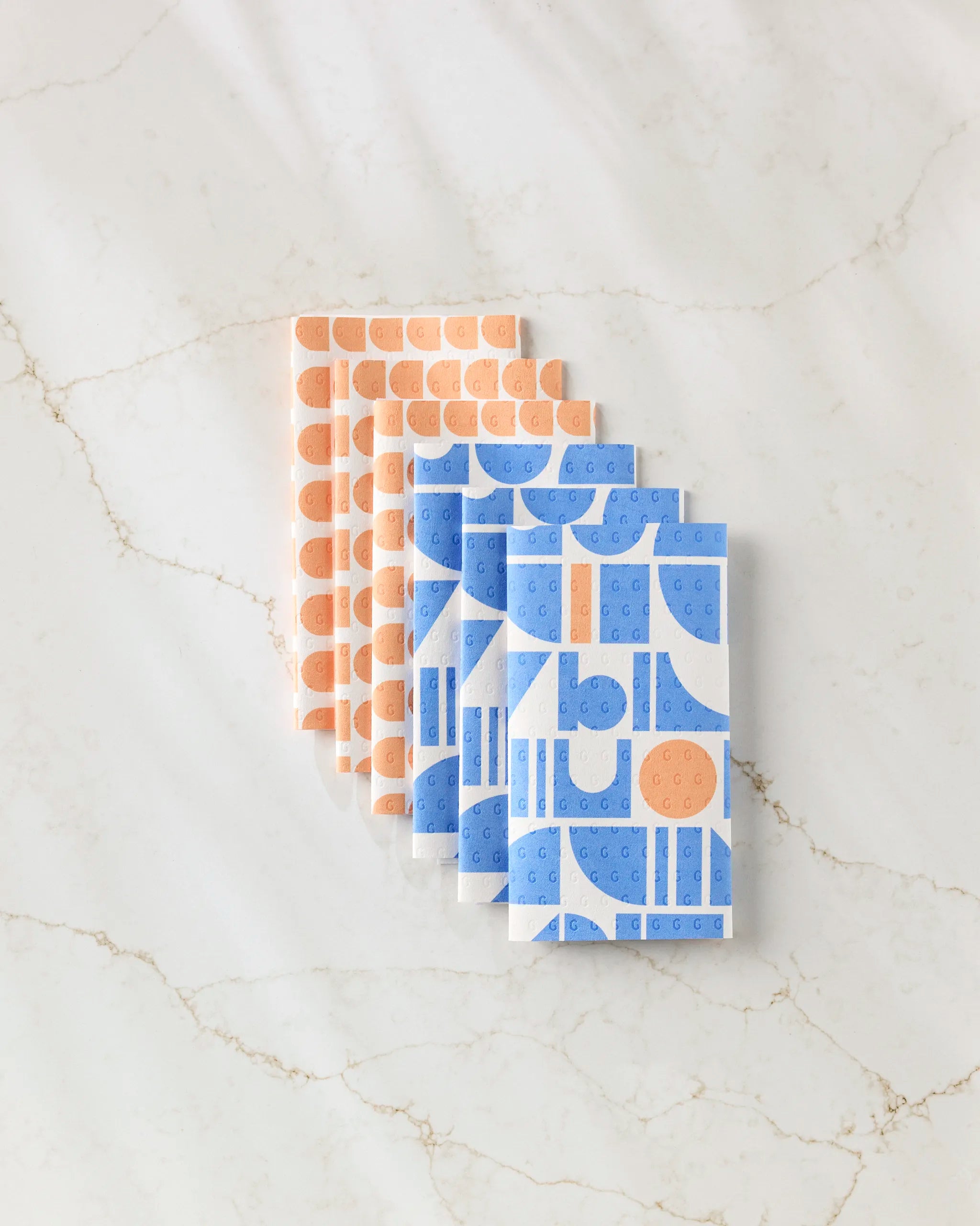 Six Sun Beam Harmony notebooks by Happy Hemera, featuring blue and peach geometric patterns on recycled paper towel covers, are neatly overlapped on a white marble surface.