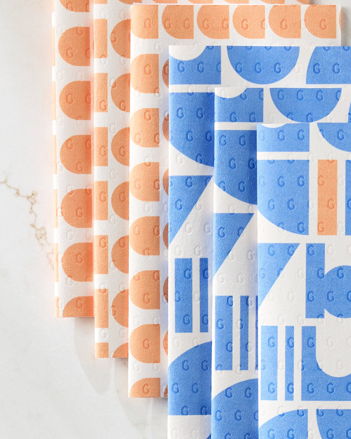 Five folded Happy Hemera Sun Beam Harmony eco-friendly kitchen towels, featuring abstract geometric patterns and the letter G, are arranged on a white marble surface—three with orange designs and two with blue.